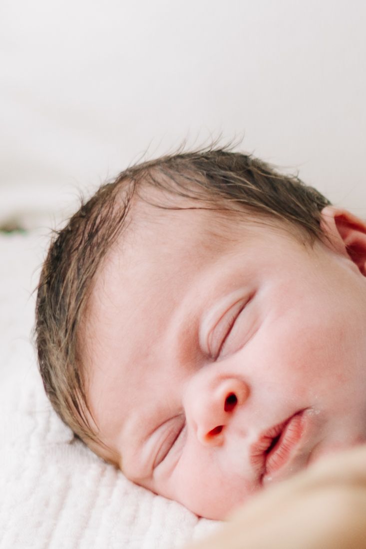 Peaceful newborn baby sleeping on a white blanket with dark hair and delicate features in a soft-lit portrait.
