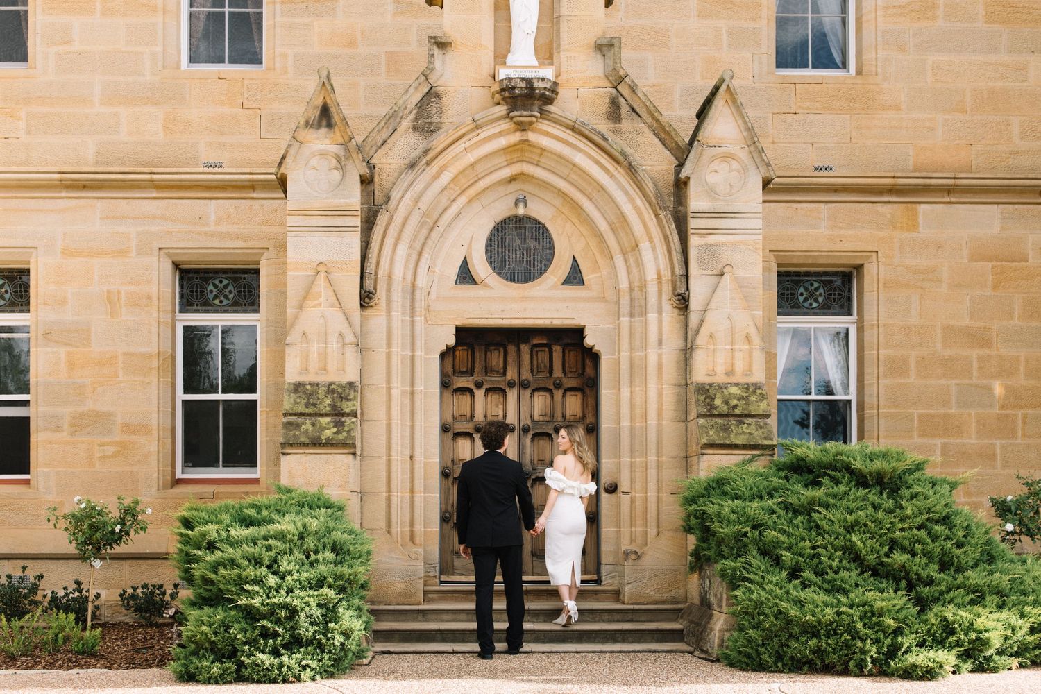 Couple walking hand in hand toward ornate stone church entrance with gothic arched doorway and lush green shrubs.