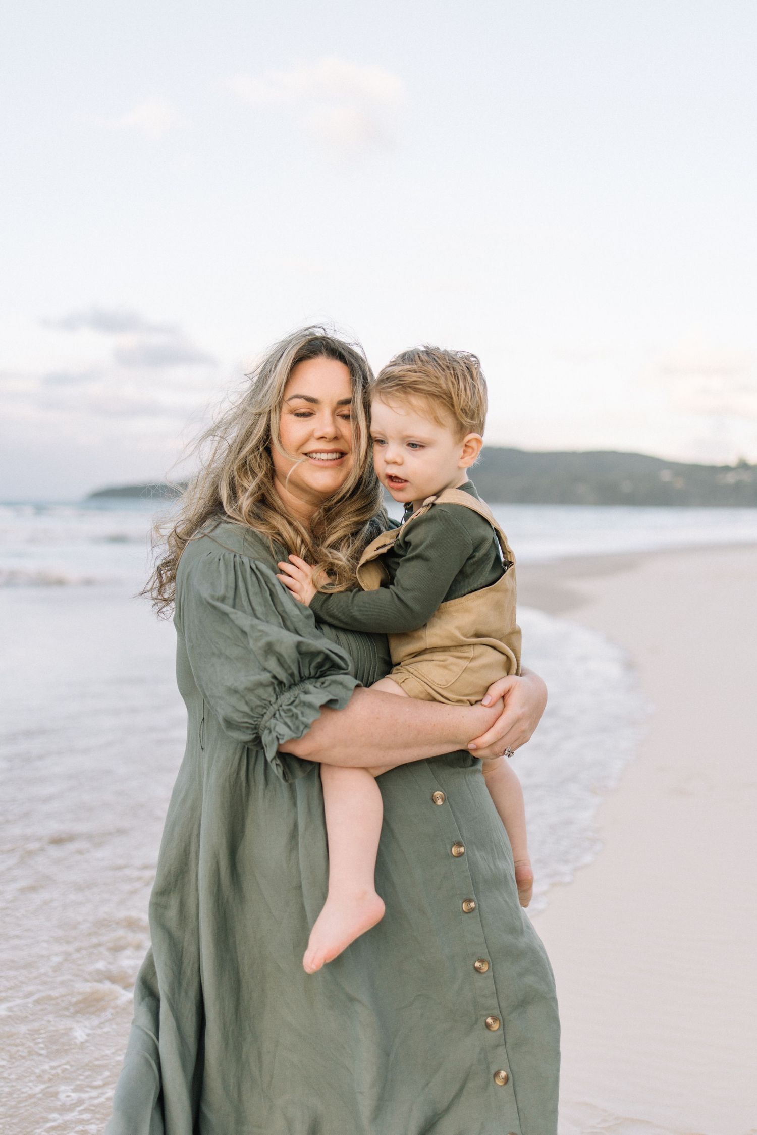 A mother in a green dress holds her toddler son on a sandy beach at sunset.