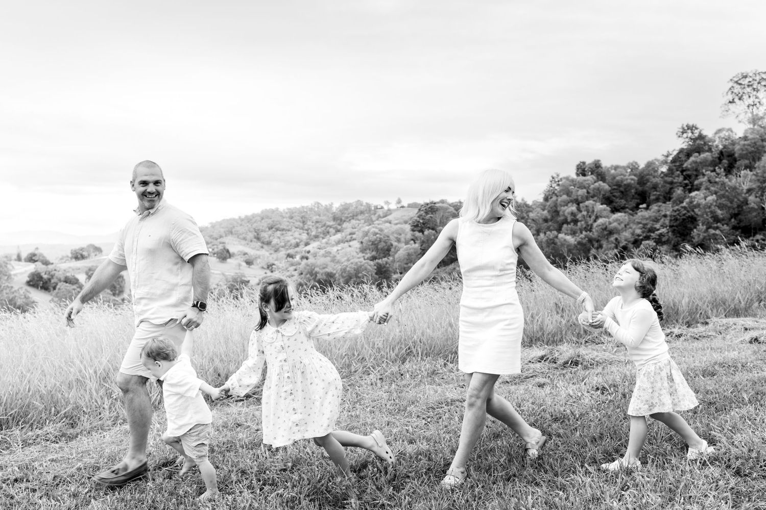 Black and white photo of a joyful family playing and holding hands in a grassy field outdoors.