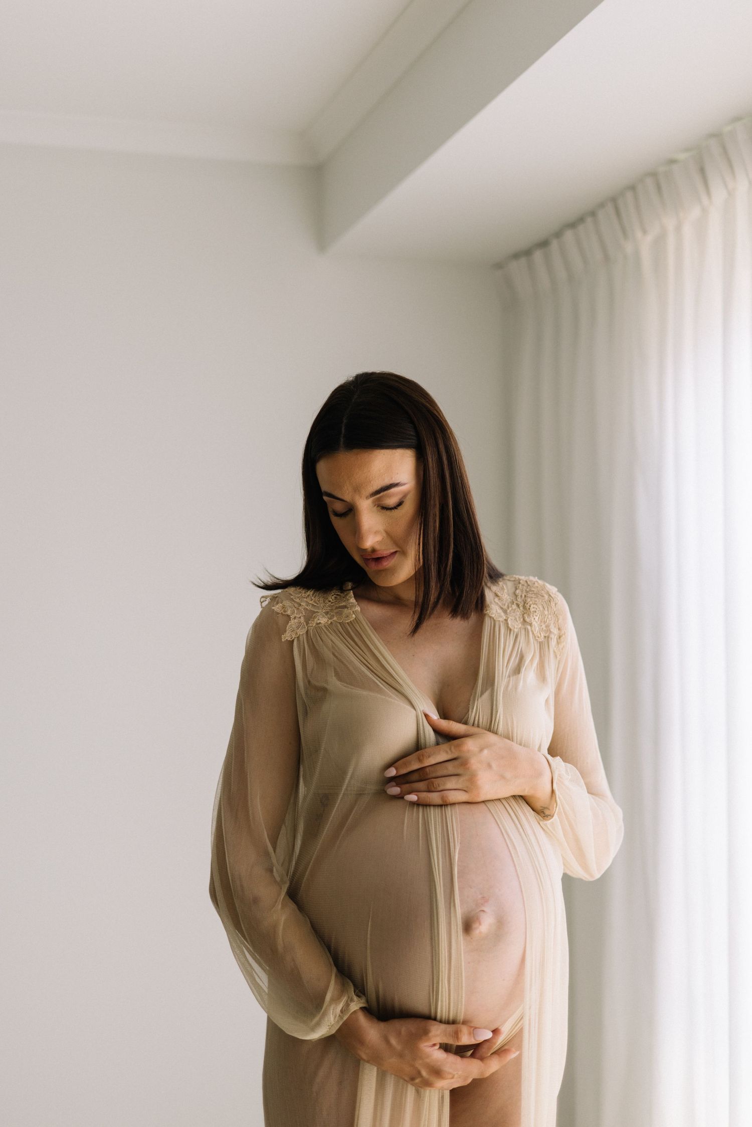 Pregnant woman in sheer beige robe tenderly cradling her baby bump while standing near a bright window.