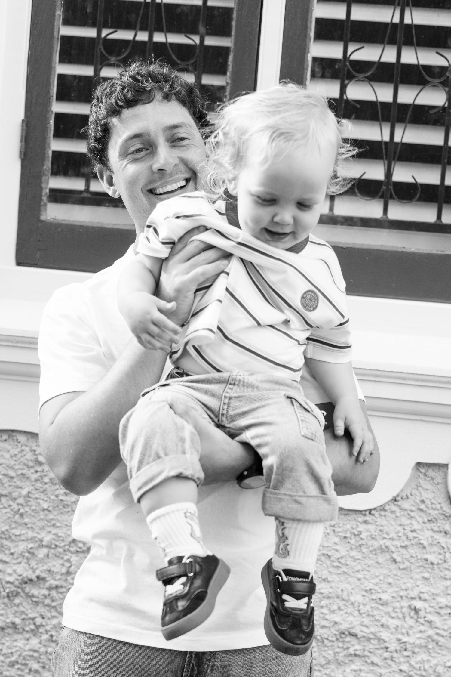 Black and white photo of a smiling man holding a happy toddler outdoors near a window.