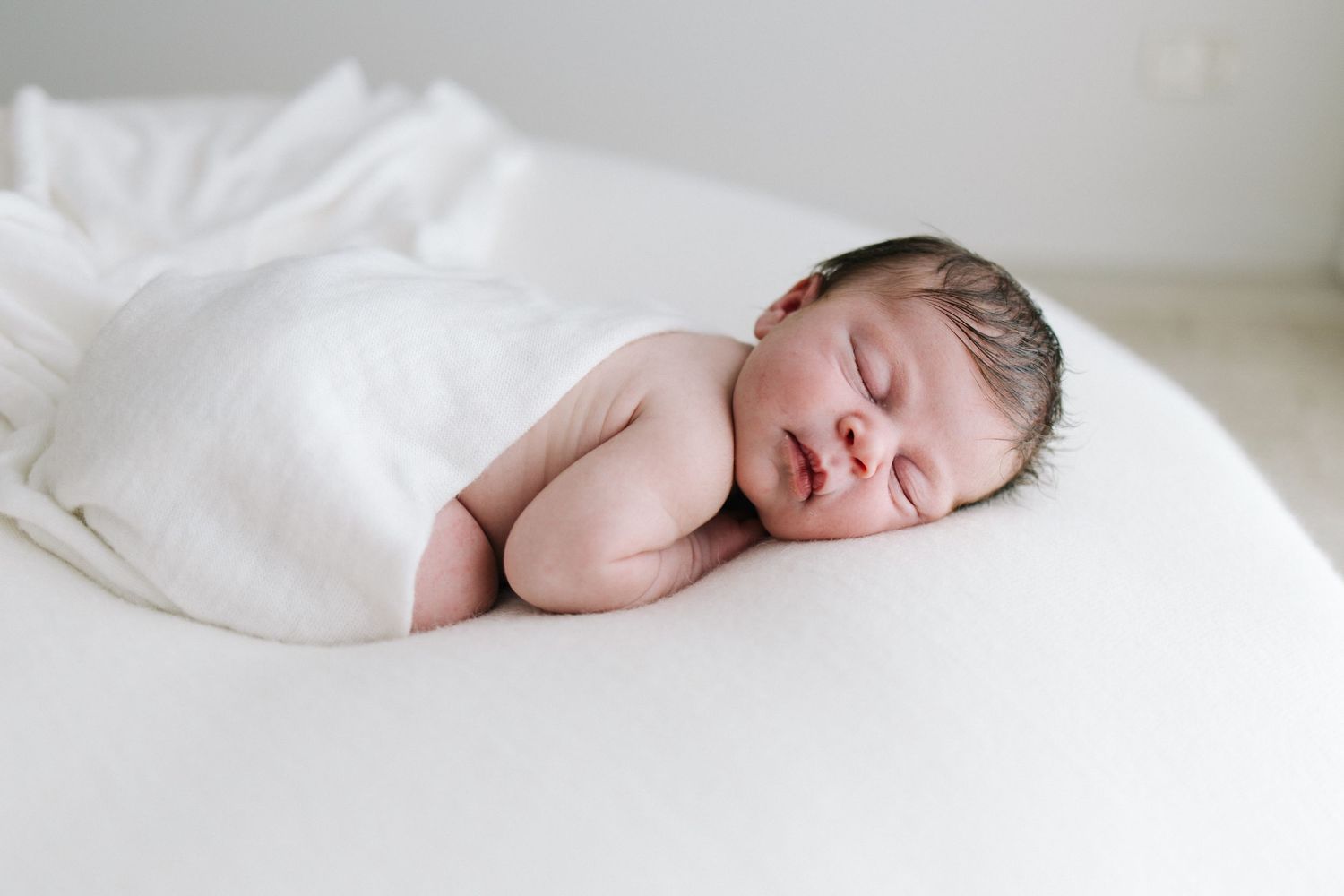 Peaceful newborn baby sleeping wrapped in white swaddle on soft white surface in bright natural light.