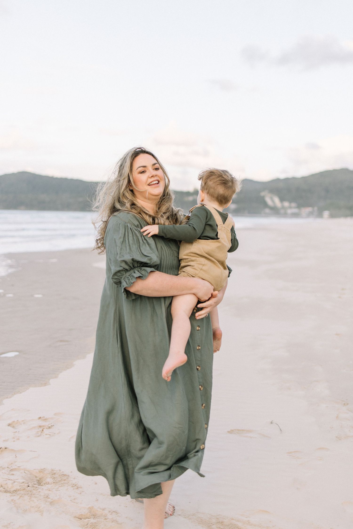 A smiling mother in a green dress holds her toddler on a sandy beach at dusk.