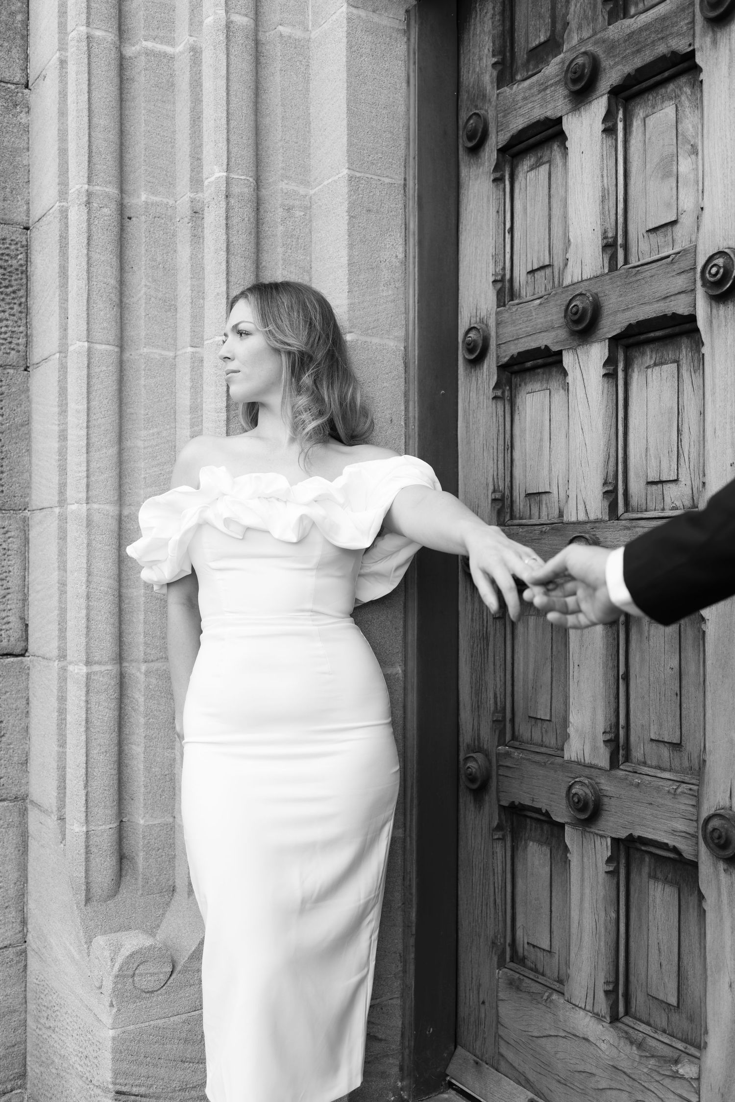 Bride in off-shoulder white dress holds groom's hand by ornate wooden church doors in black and white photo.