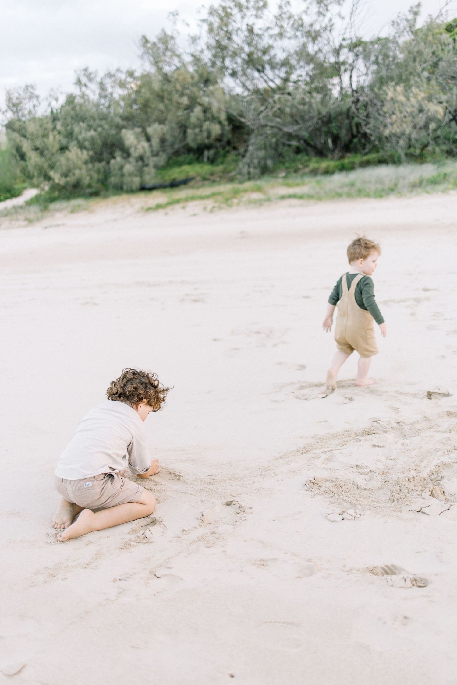 Two young children playing in white sand on a beach, one crouching and drawing in sand, the other toddling nearby.
