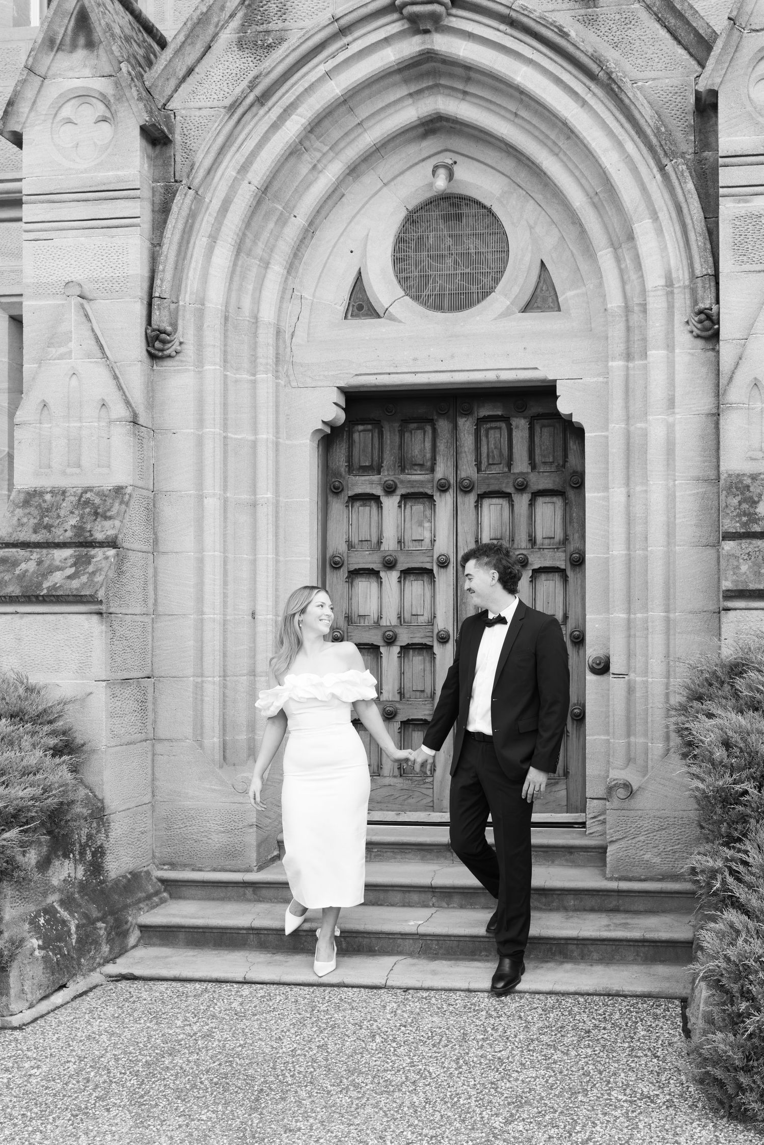 Black and white photo of an elegant couple holding hands outside a stone church doorway.