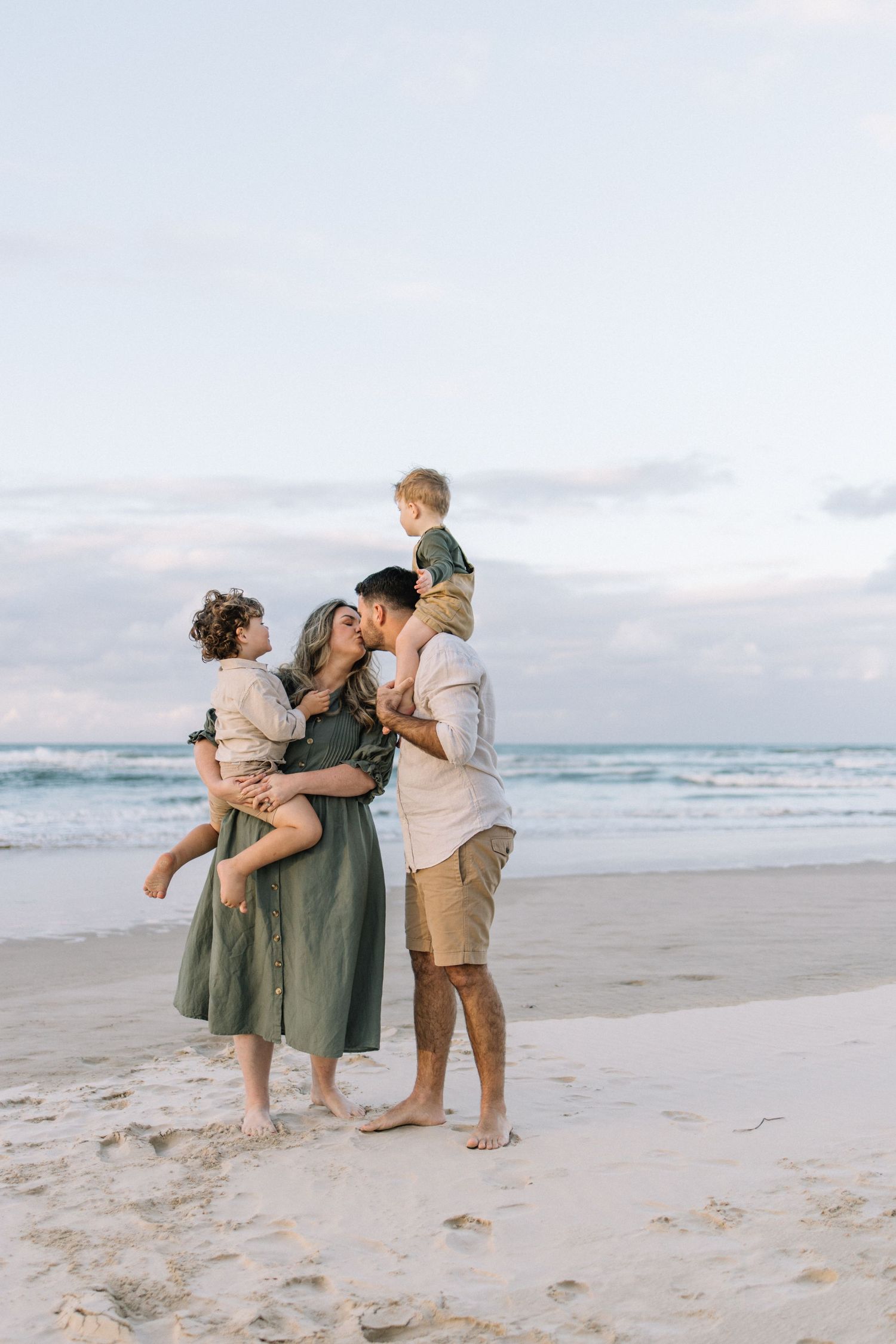 Family of four sharing a tender kiss on a sandy beach at sunset, parents holding young children.