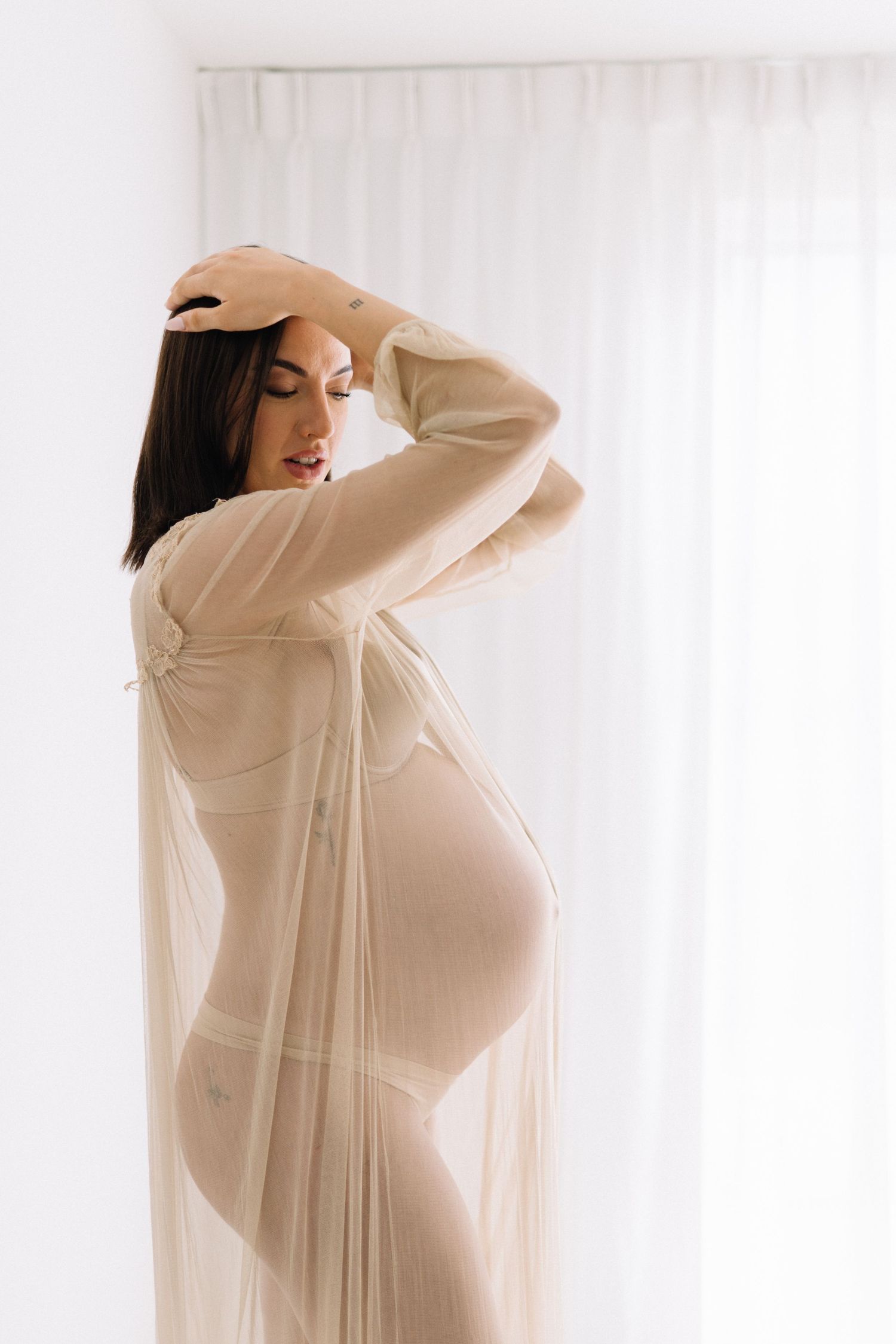 Pregnant woman in sheer beige robe posing gracefully near white curtains in a soft, elegant maternity photo.
