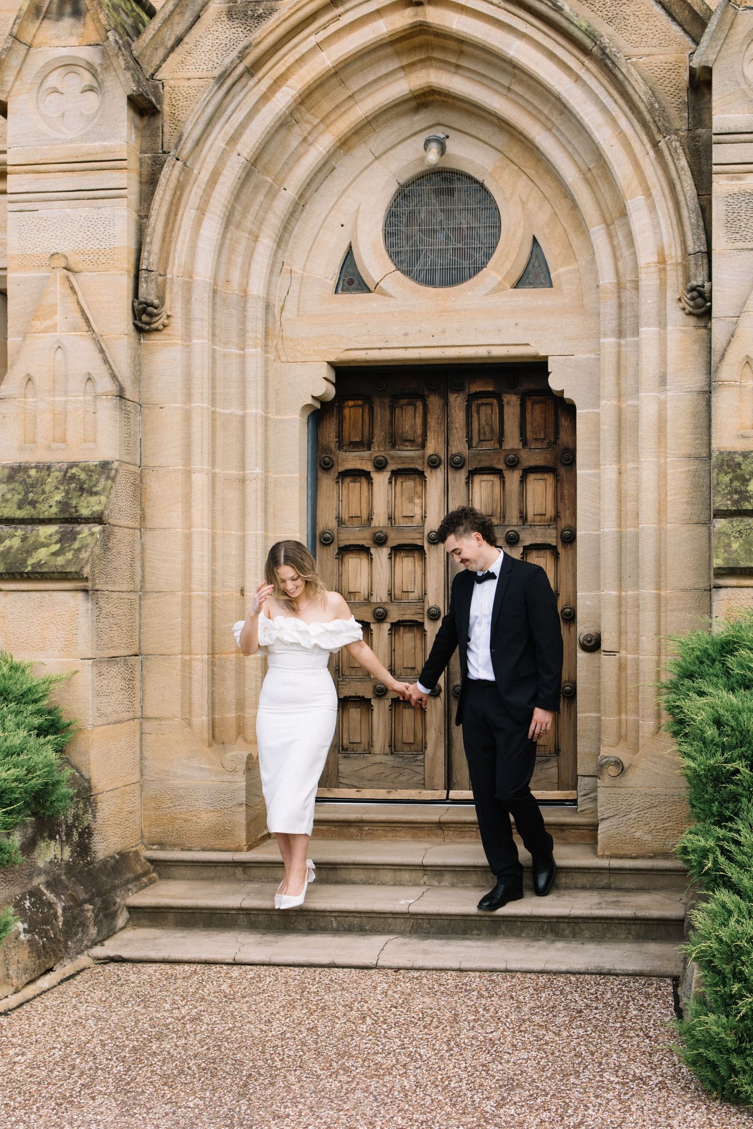 Bride and groom holding hands on church steps, she in white off-shoulder dress, he in black tuxedo with bow tie.