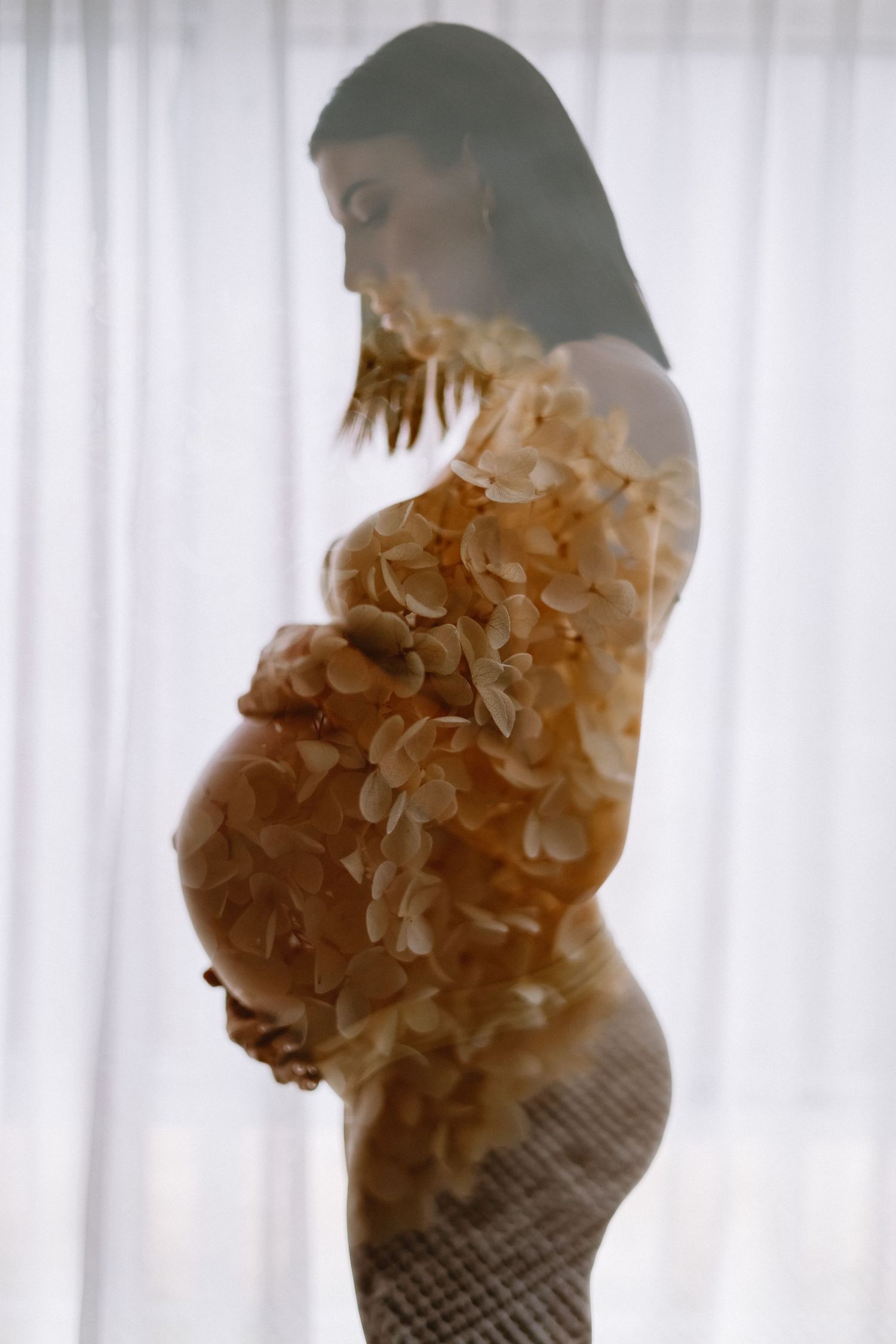 Pregnant woman in profile wearing golden floral arrangement, double exposure maternity portrait by sheer curtains.