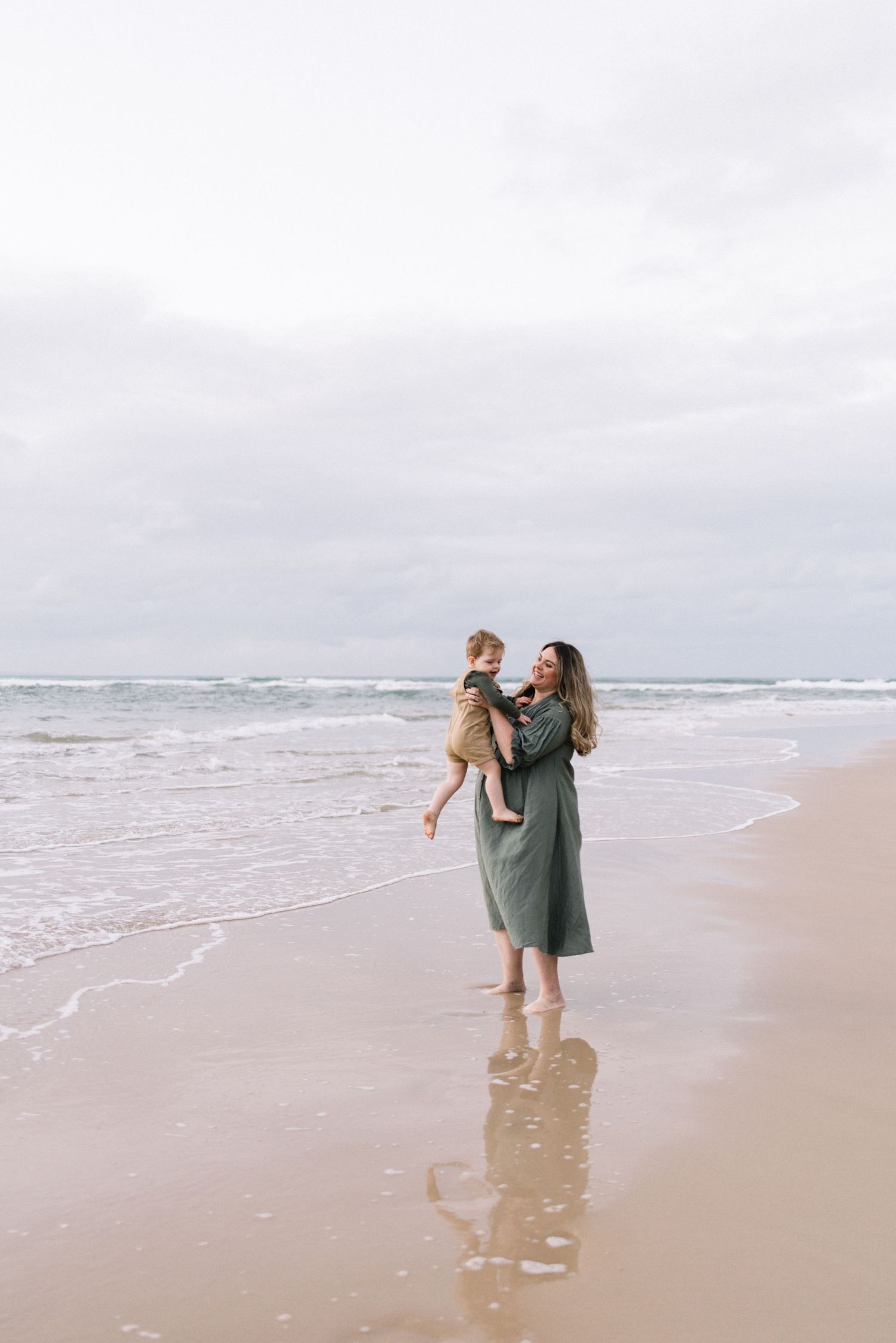 A mother in a green maxi dress lifts her smiling toddler near ocean waves on a beach.