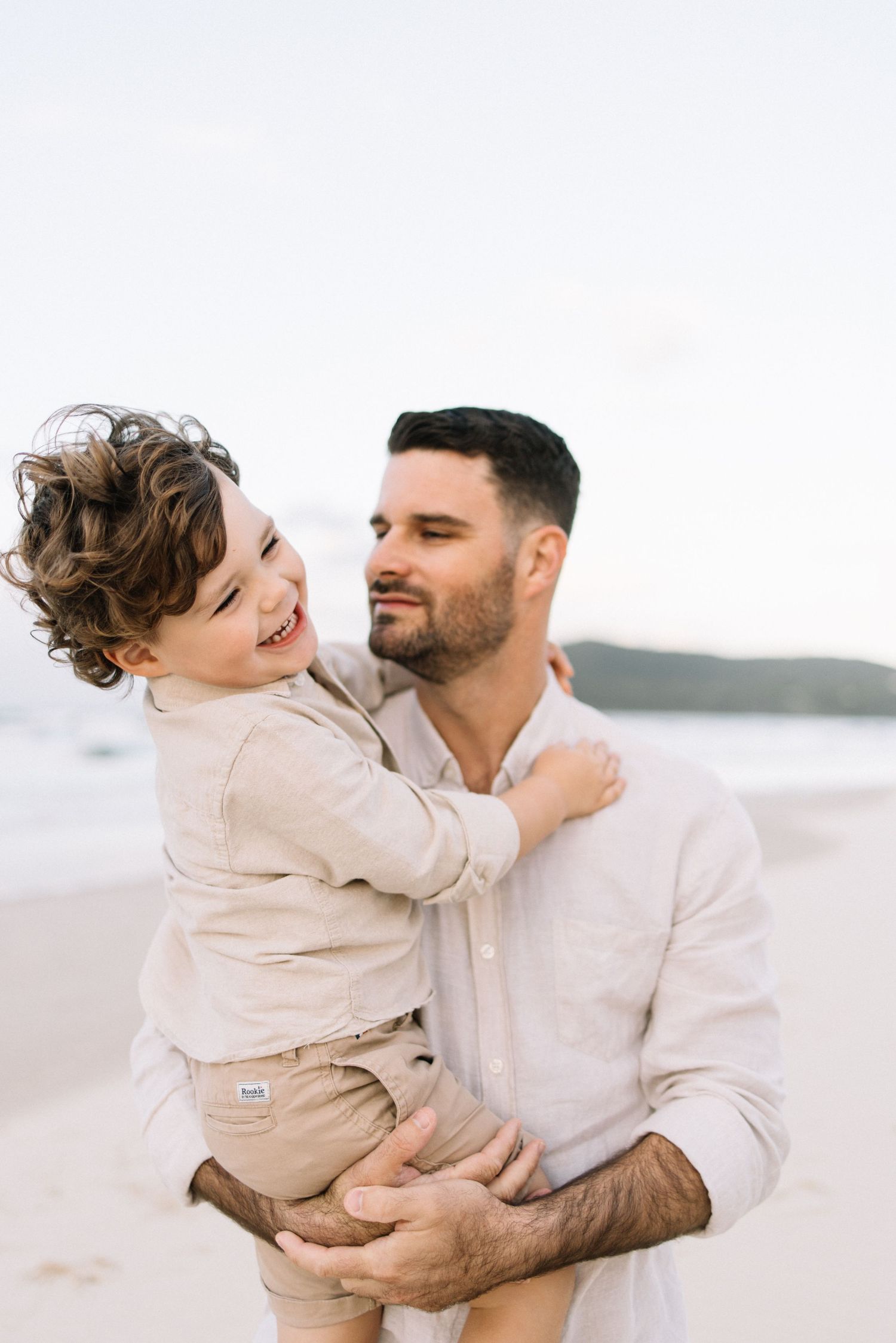 A father holds his laughing curly-haired son on a sandy beach, sharing a joyful moment together.