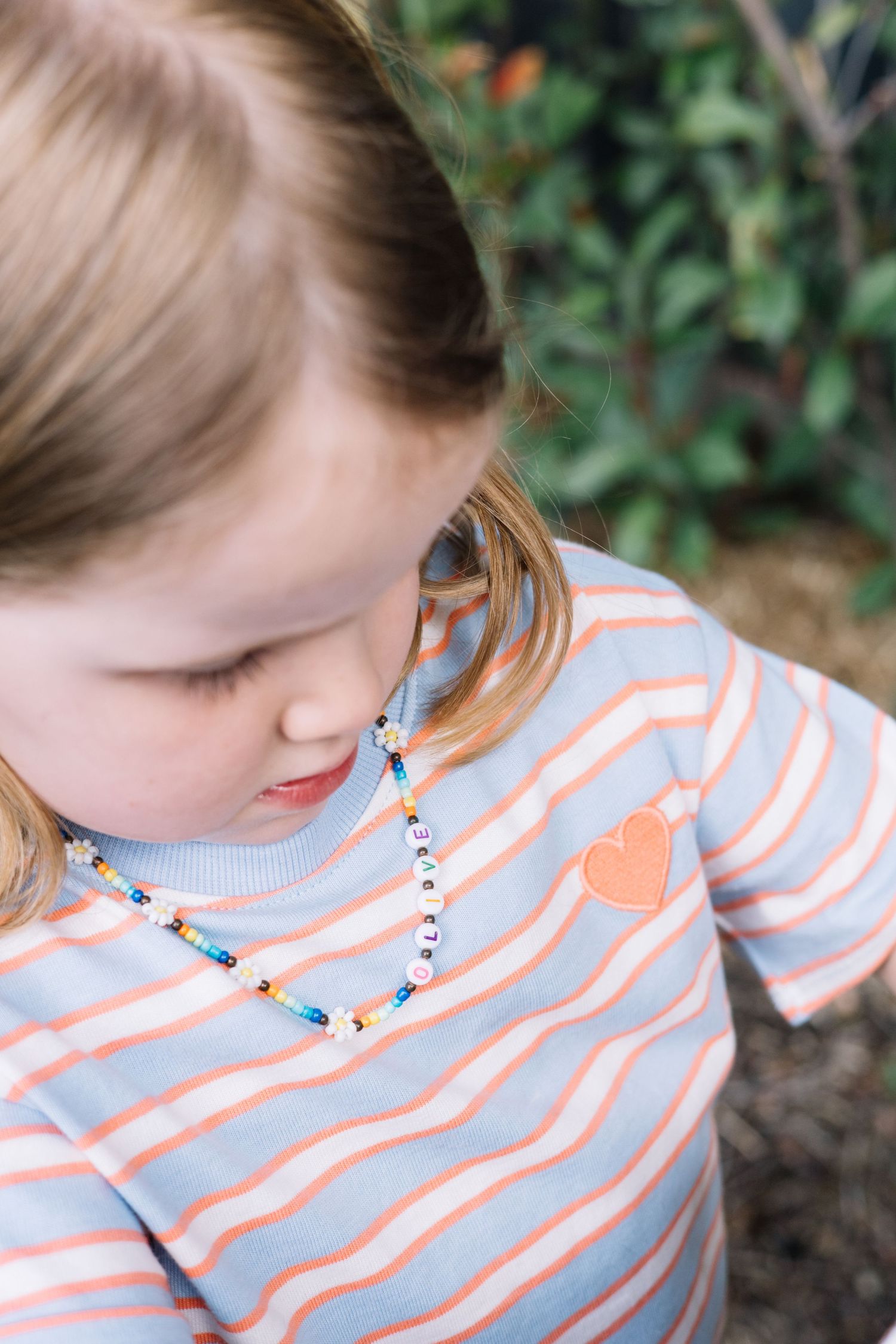 Young girl wearing a striped blue and coral shirt with heart patch and colorful beaded necklace outdoors.