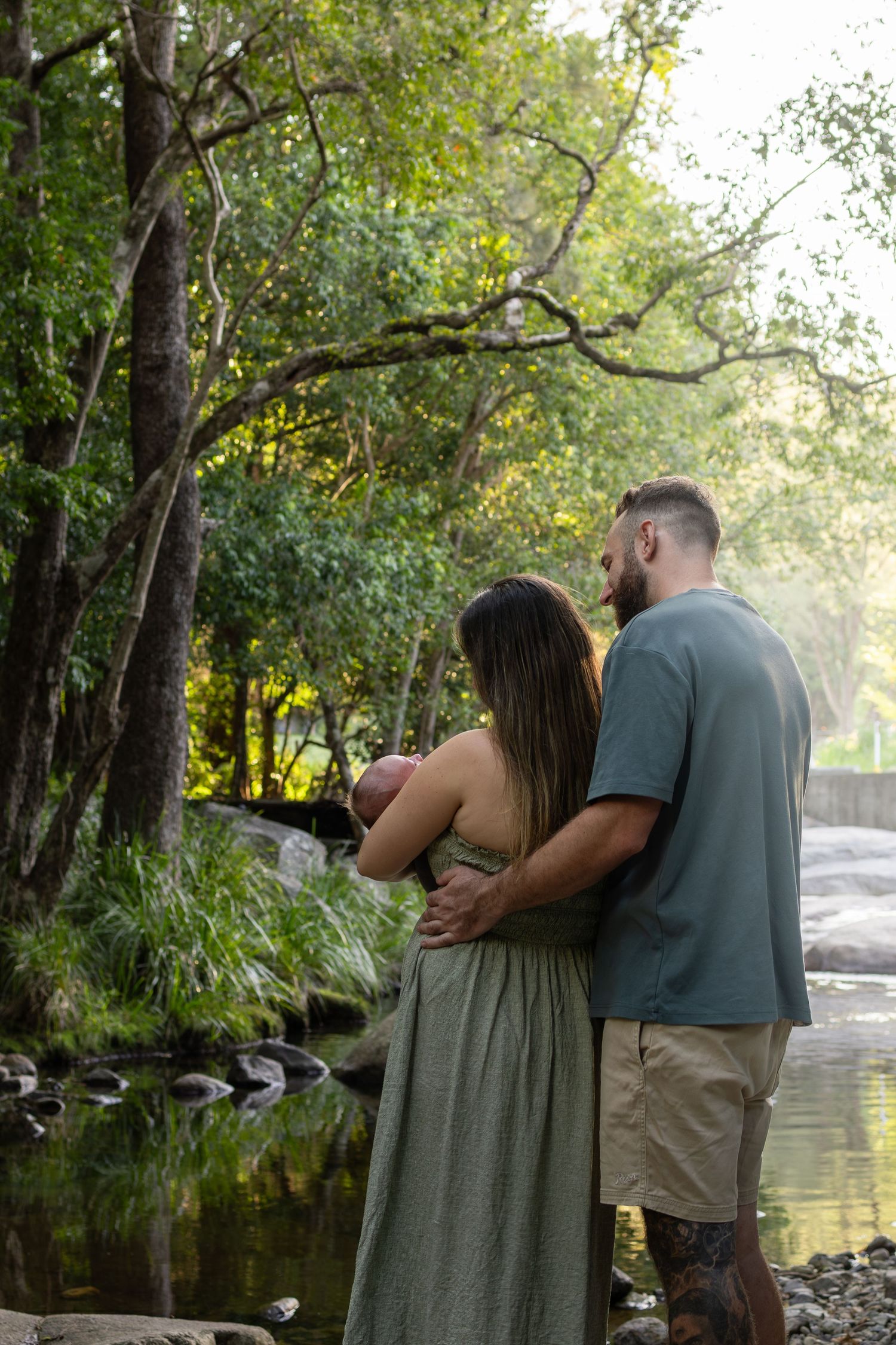 Couple embracing by a serene forest creek, surrounded by lush green trees in soft natural light.