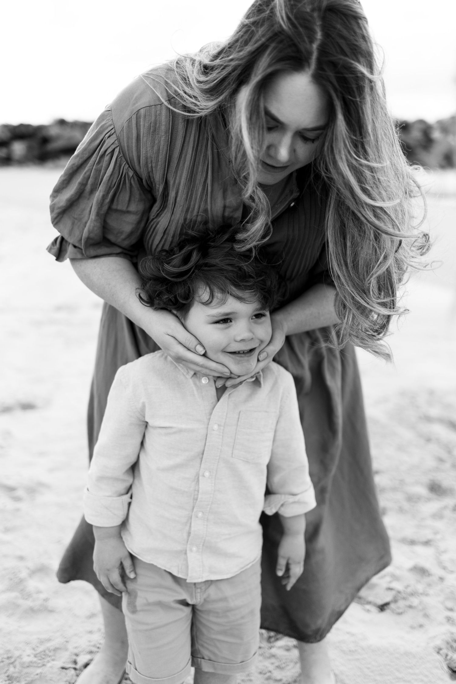 Black and white photo of a mother with long hair hugging her curly-haired toddler son on a sandy beach.