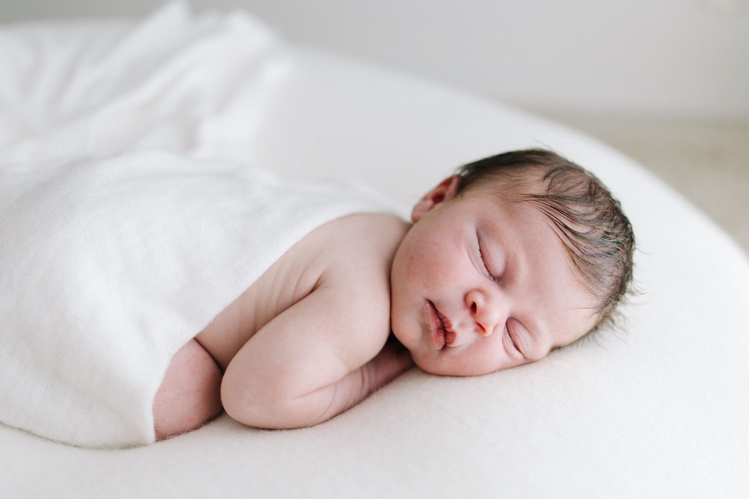 Peaceful newborn baby sleeping on white surface, wrapped in soft white blanket, newborn photography.