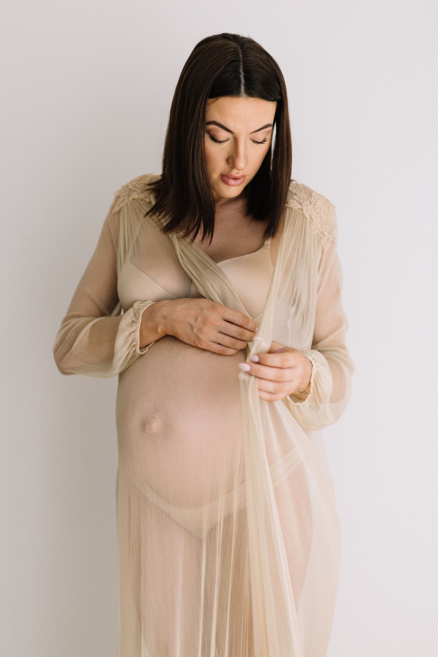 Pregnant woman in sheer beige maternity gown cradling her baby bump against a white background.