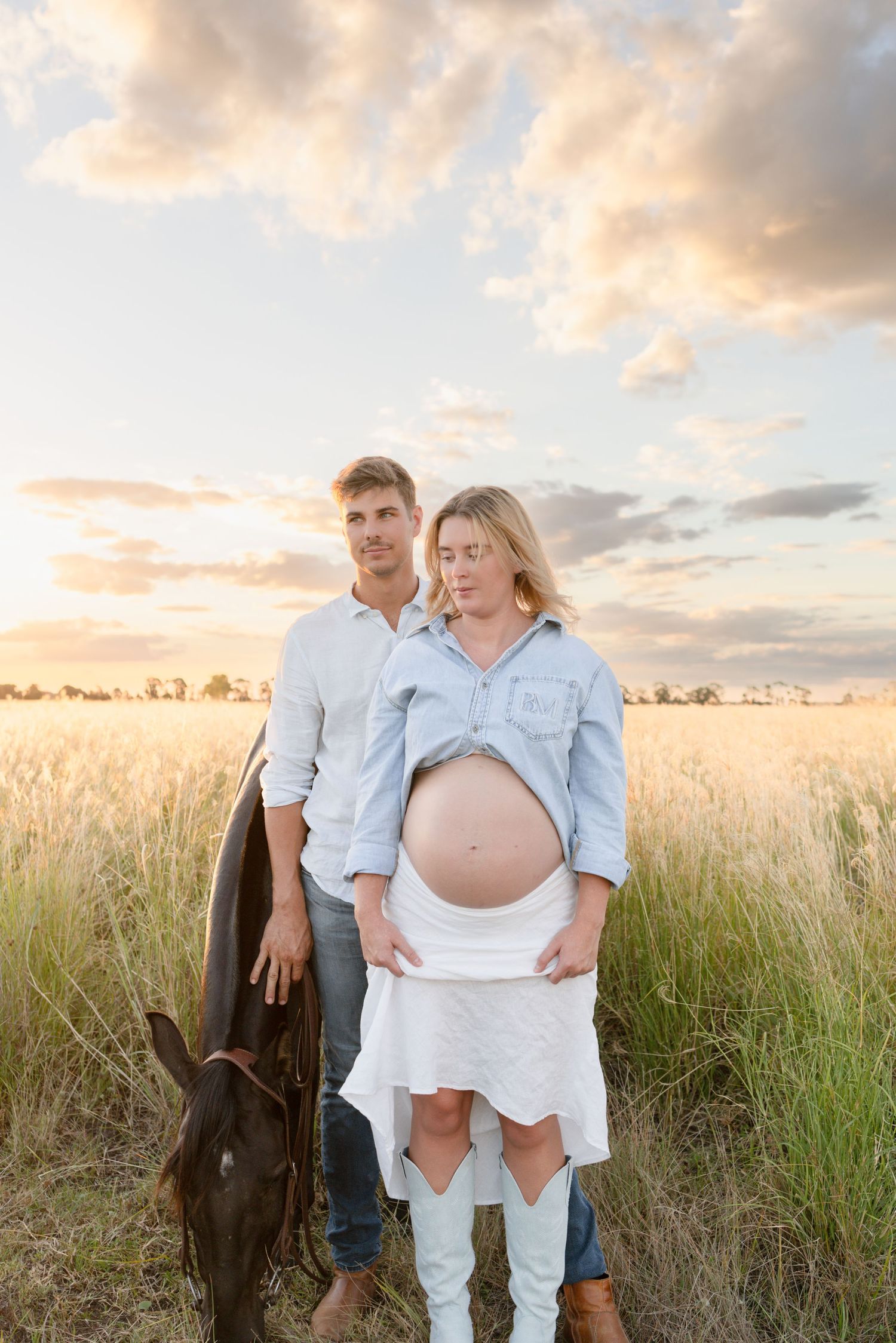 Couple standing in golden field at sunset, woman showing pregnant belly.