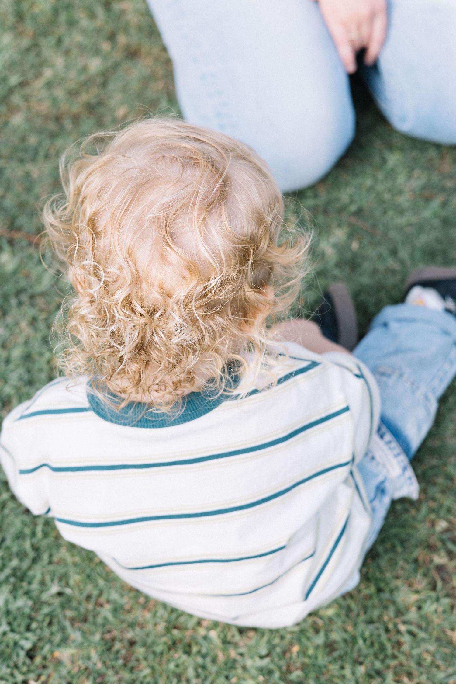 Toddler with curly blonde hair sits on green grass wearing a striped shirt and jeans, viewed from behind.