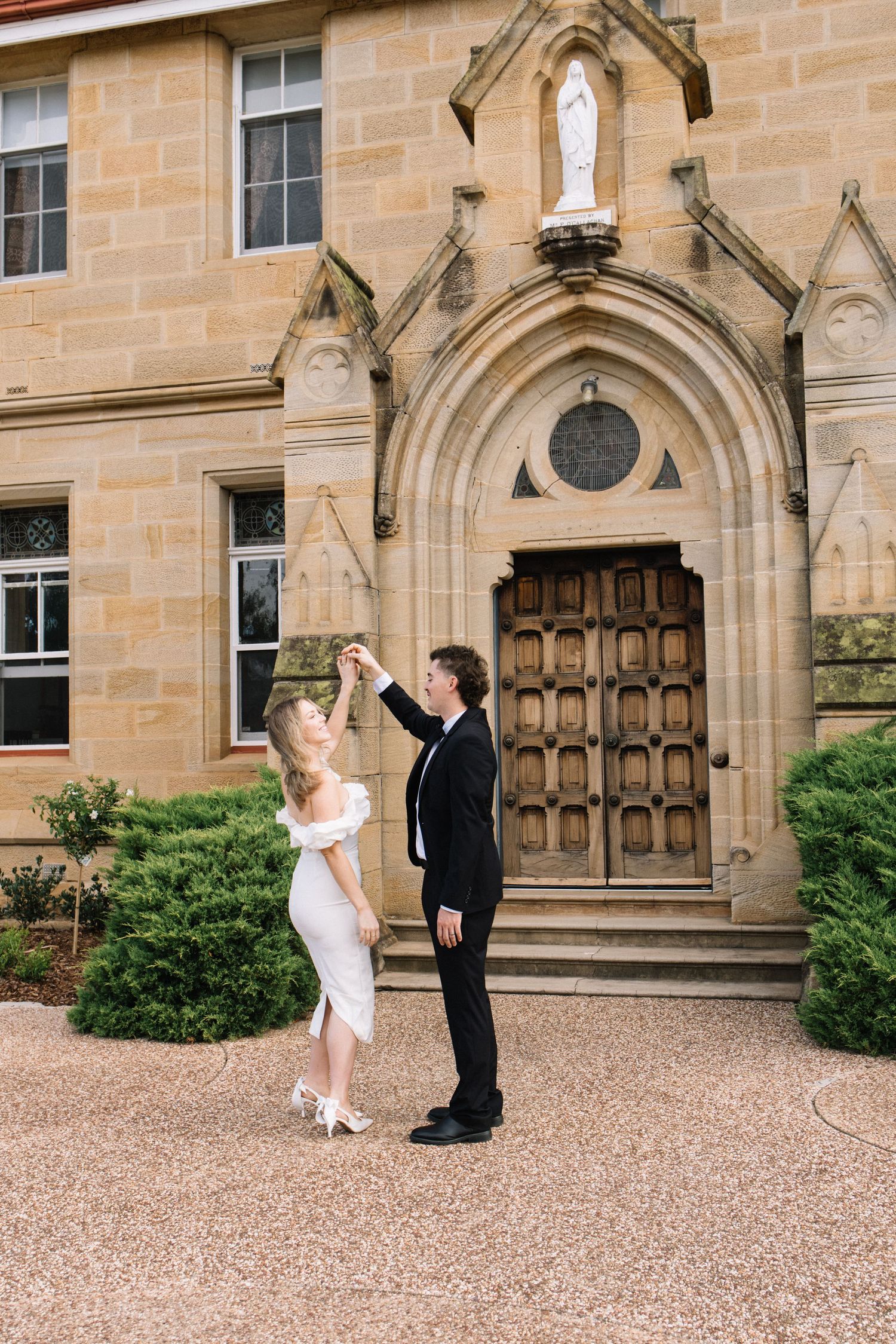 Bride and groom dancing outside a historic stone church with ornate wooden doors and a Madonna statue.