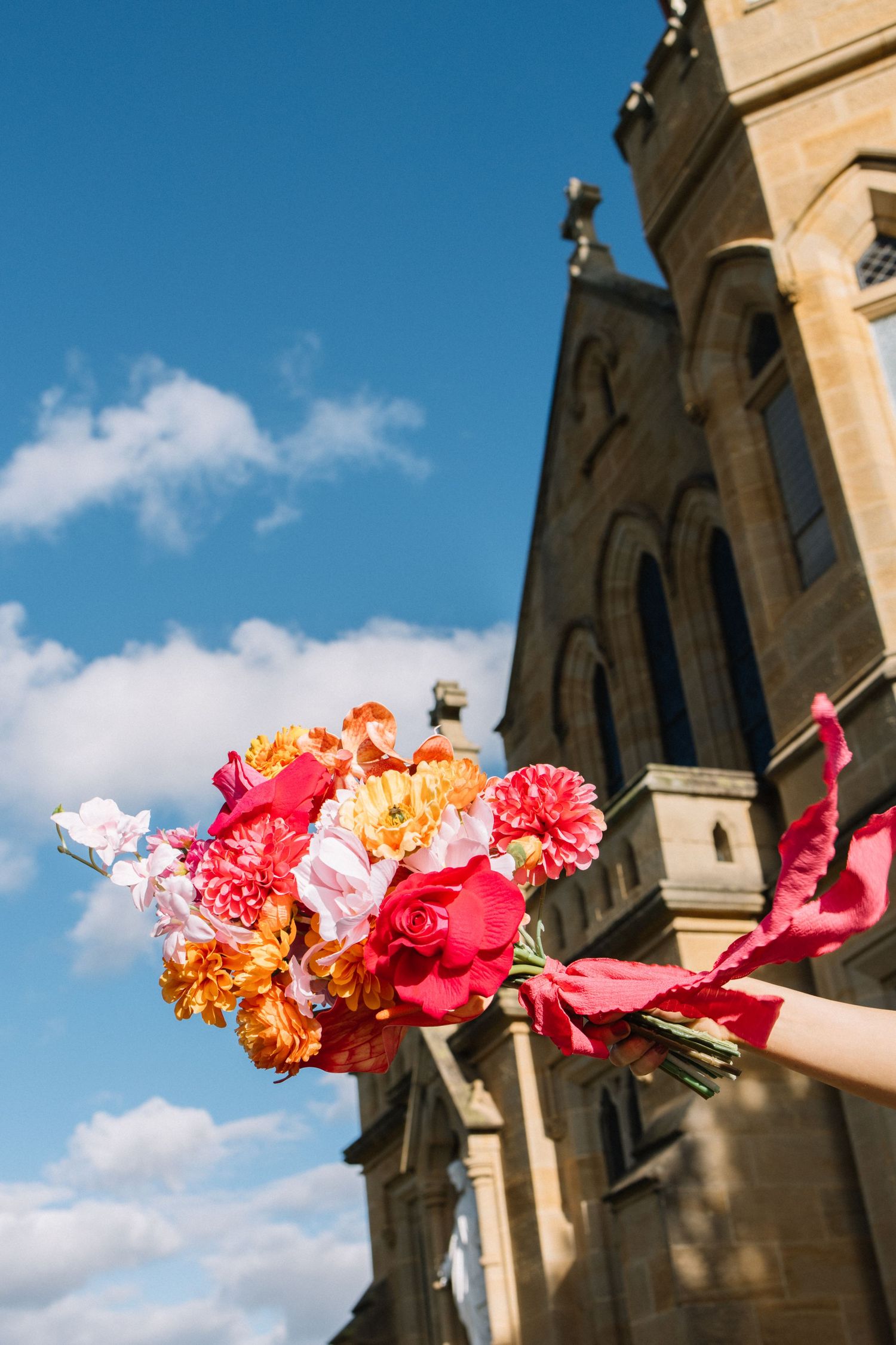 A vibrant bridal bouquet of red, pink, and orange flowers held up against a blue sky with a historic stone church.
