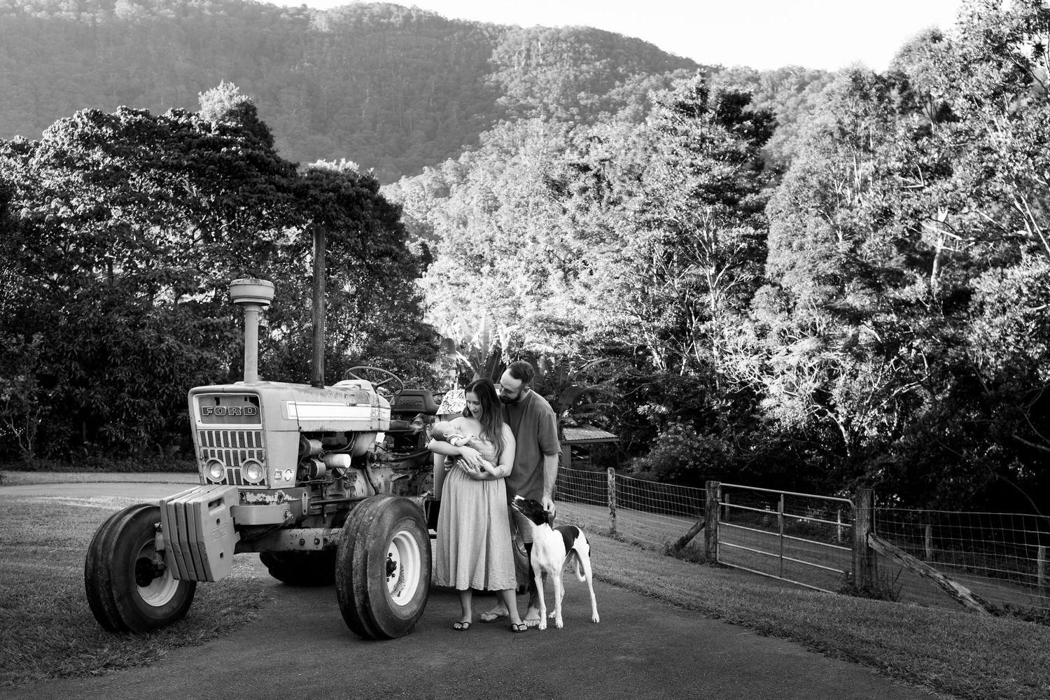 Black and white photo of a couple with a dog standing beside a vintage tractor on a rural farm with lush green hills.
