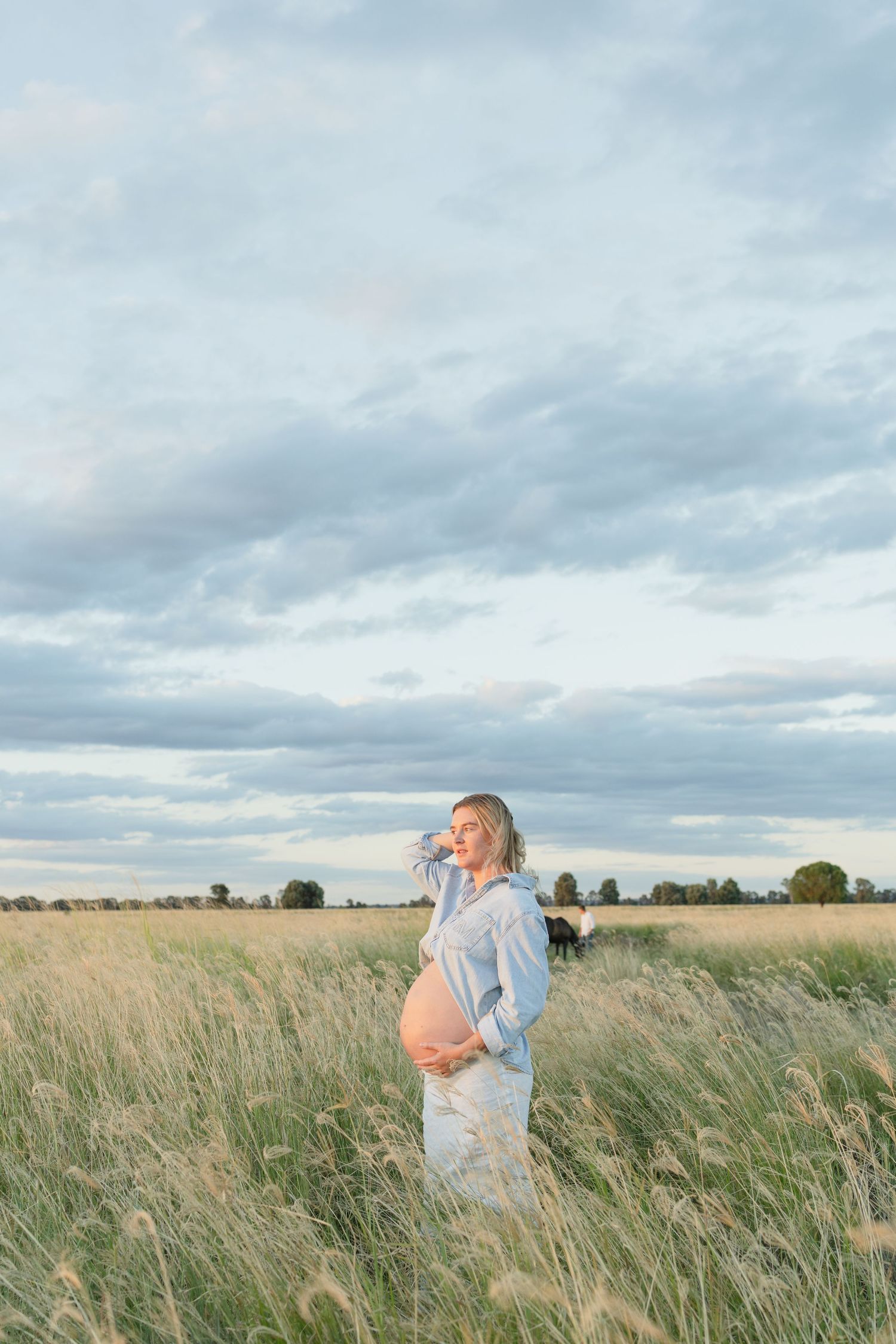 Pregnant blonde woman holding belly in tall grass field under cloudy sky.