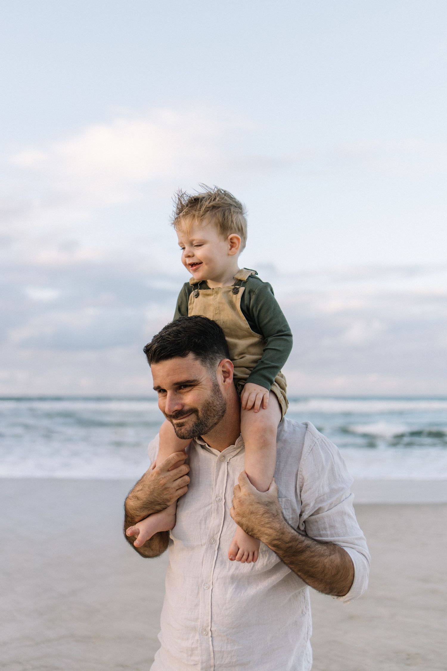 A smiling father carries his happy toddler son on his shoulders at the beach during golden hour.
