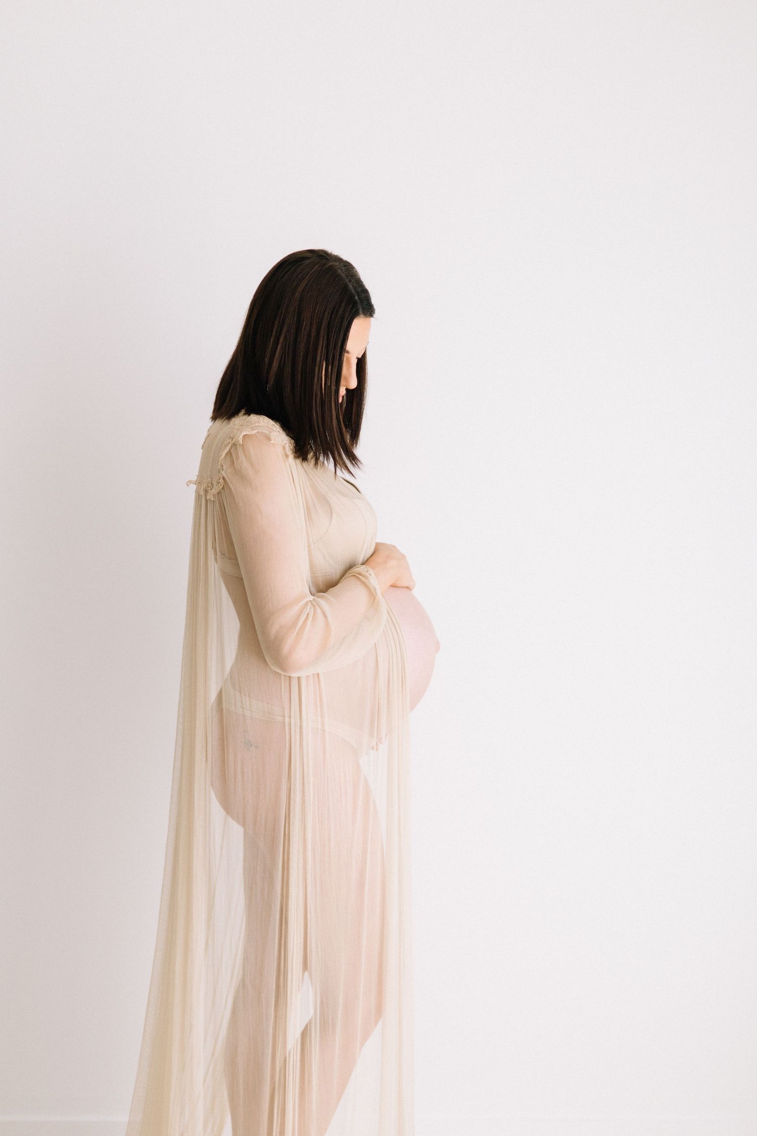 Side profile of pregnant brunette woman in sheer nude maternity gown looking down at her belly.