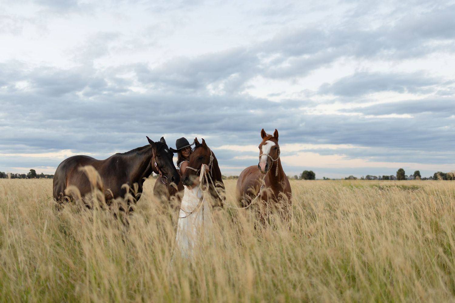 Woman in white dress and cowboy hat stands among three horses in a vast golden grass field.