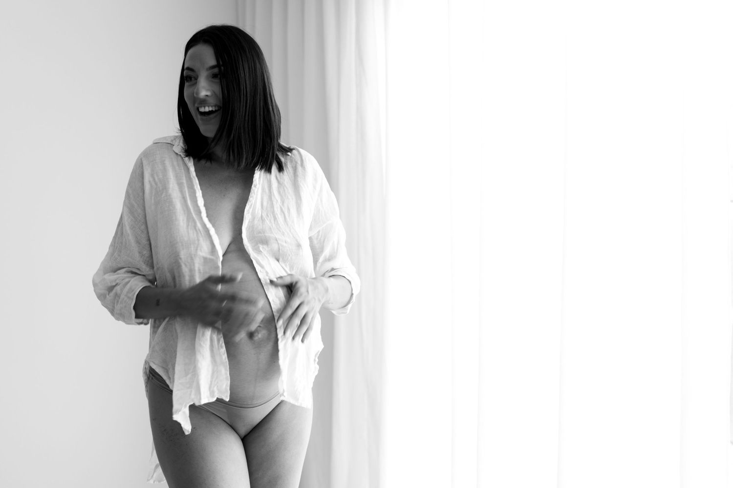 Black and white photo of a laughing pregnant woman in white shirt and underwear.