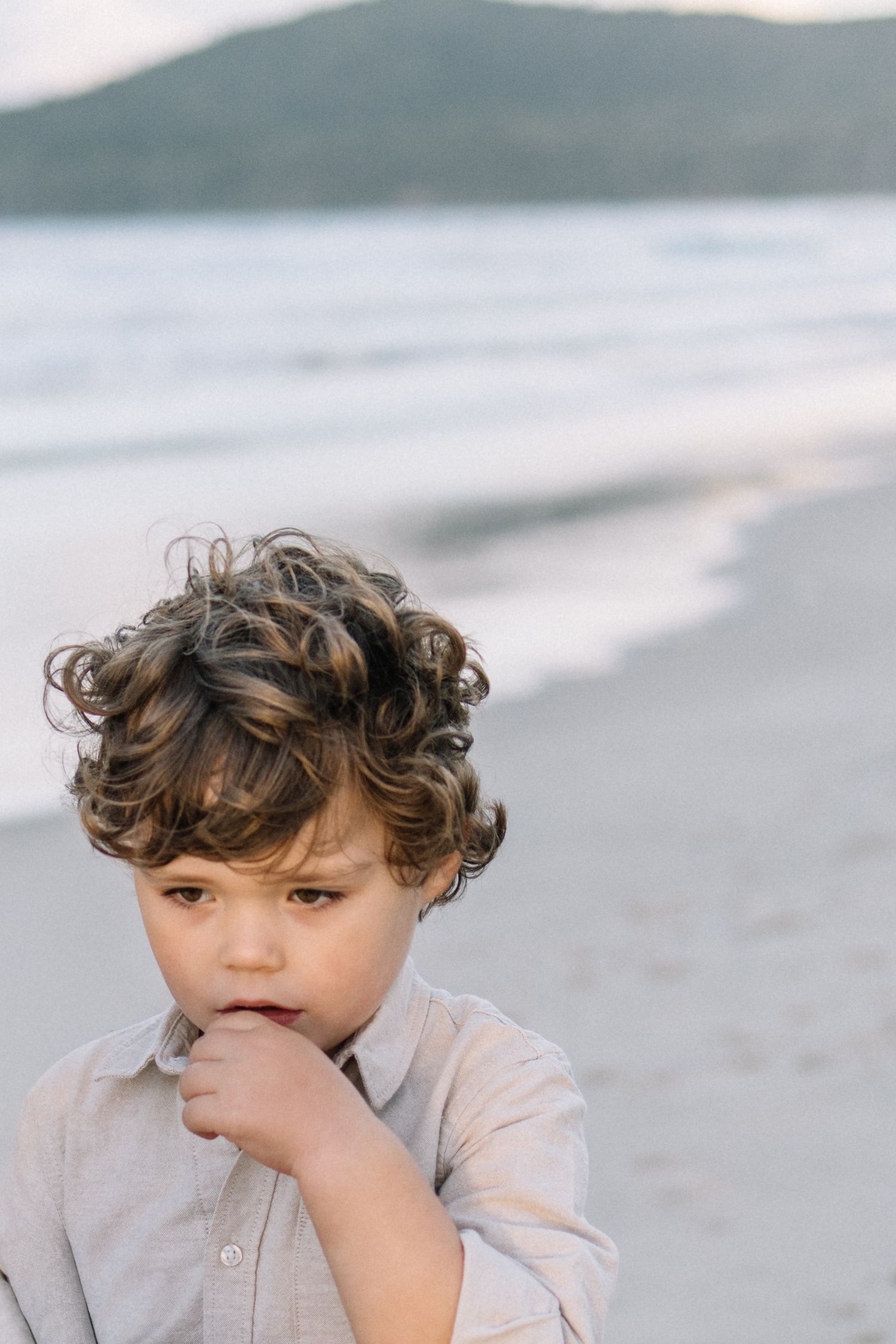 Curly-haired young boy in a linen shirt standing thoughtfully on a sandy beach with ocean waves behind him.