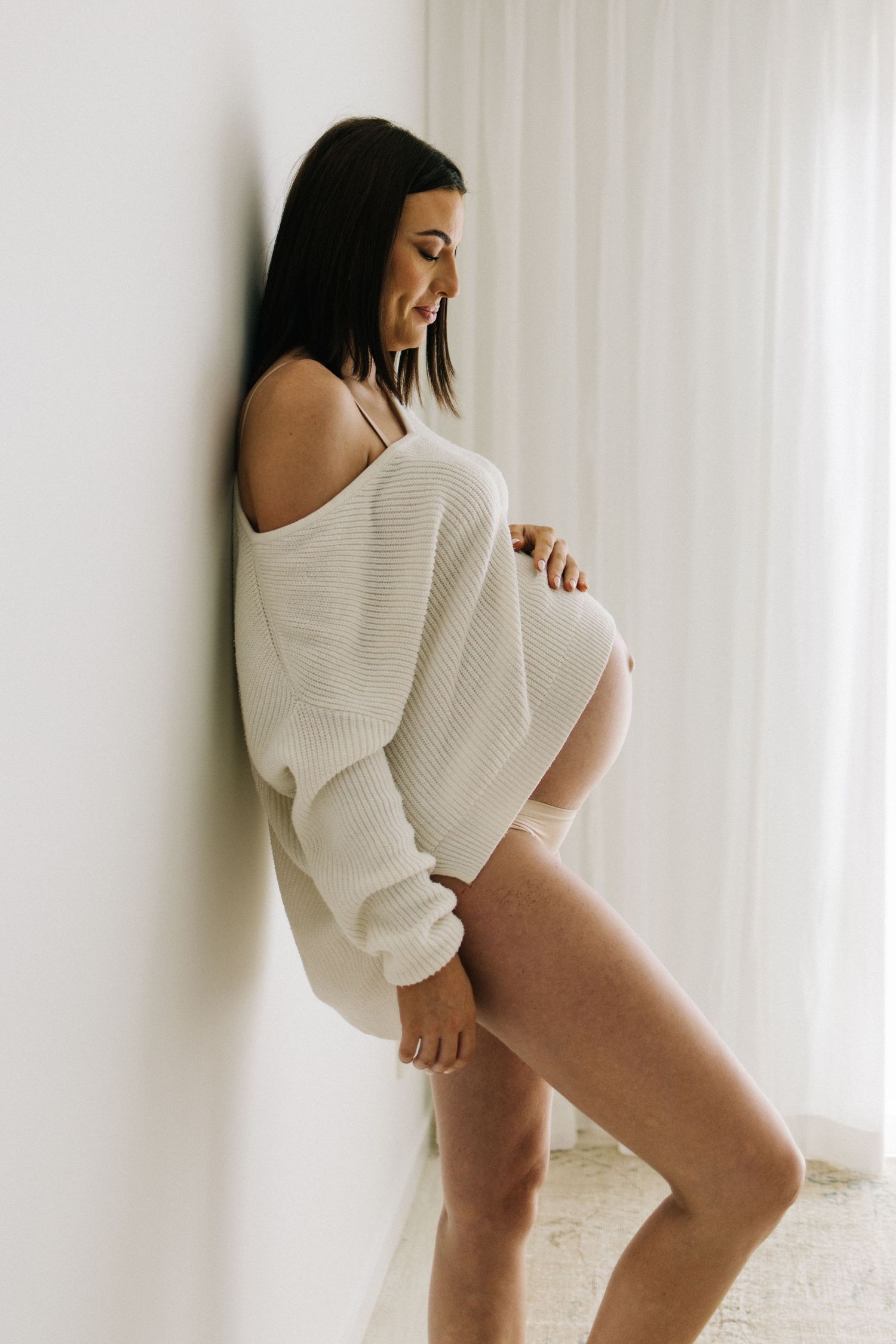 Pregnant woman in white off-shoulder sweater and beige underwear cradling baby bump against white wall.