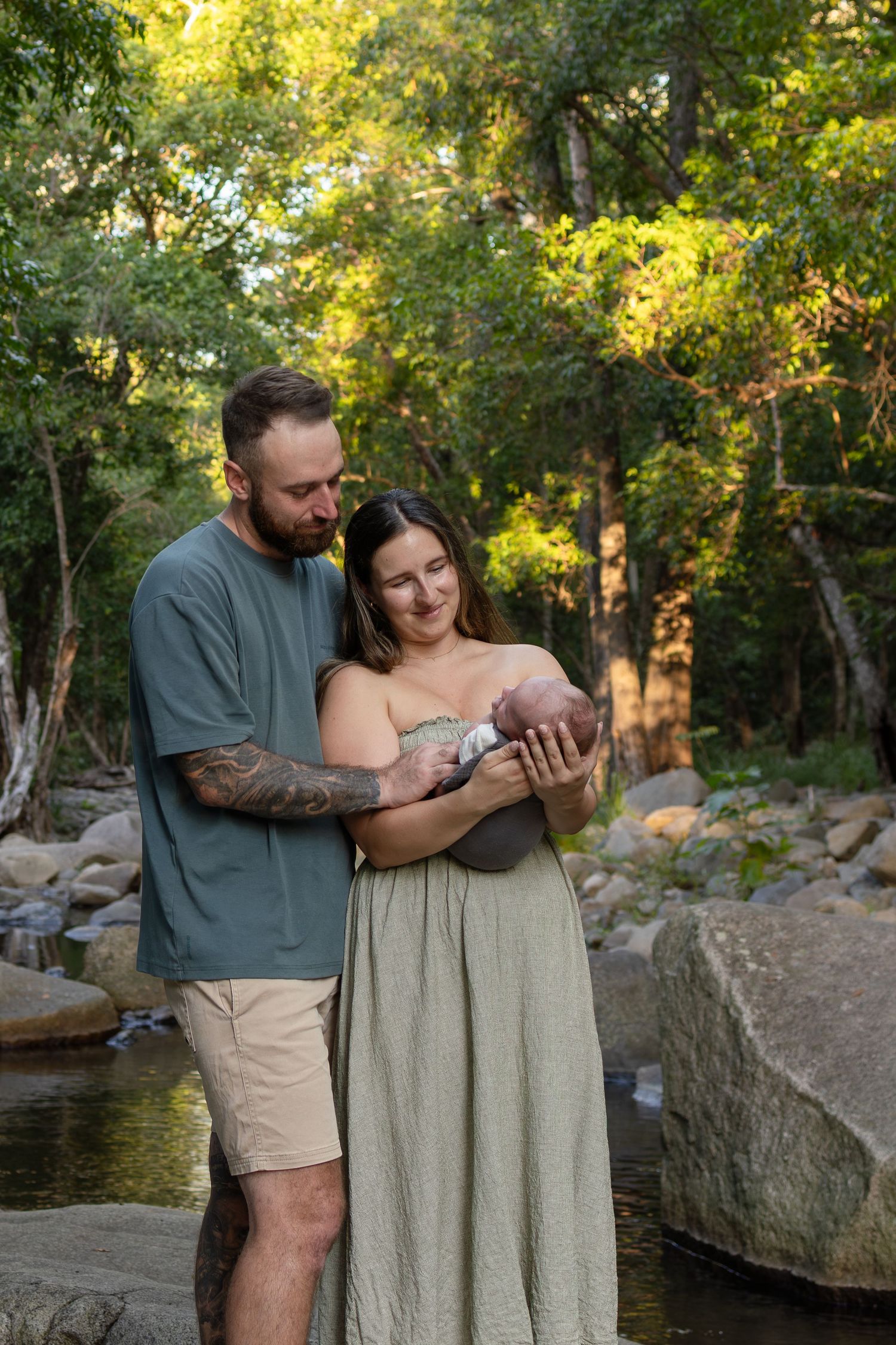 A couple stands by a rocky creek in a forest, lovingly gazing at their newborn baby in soft golden light.