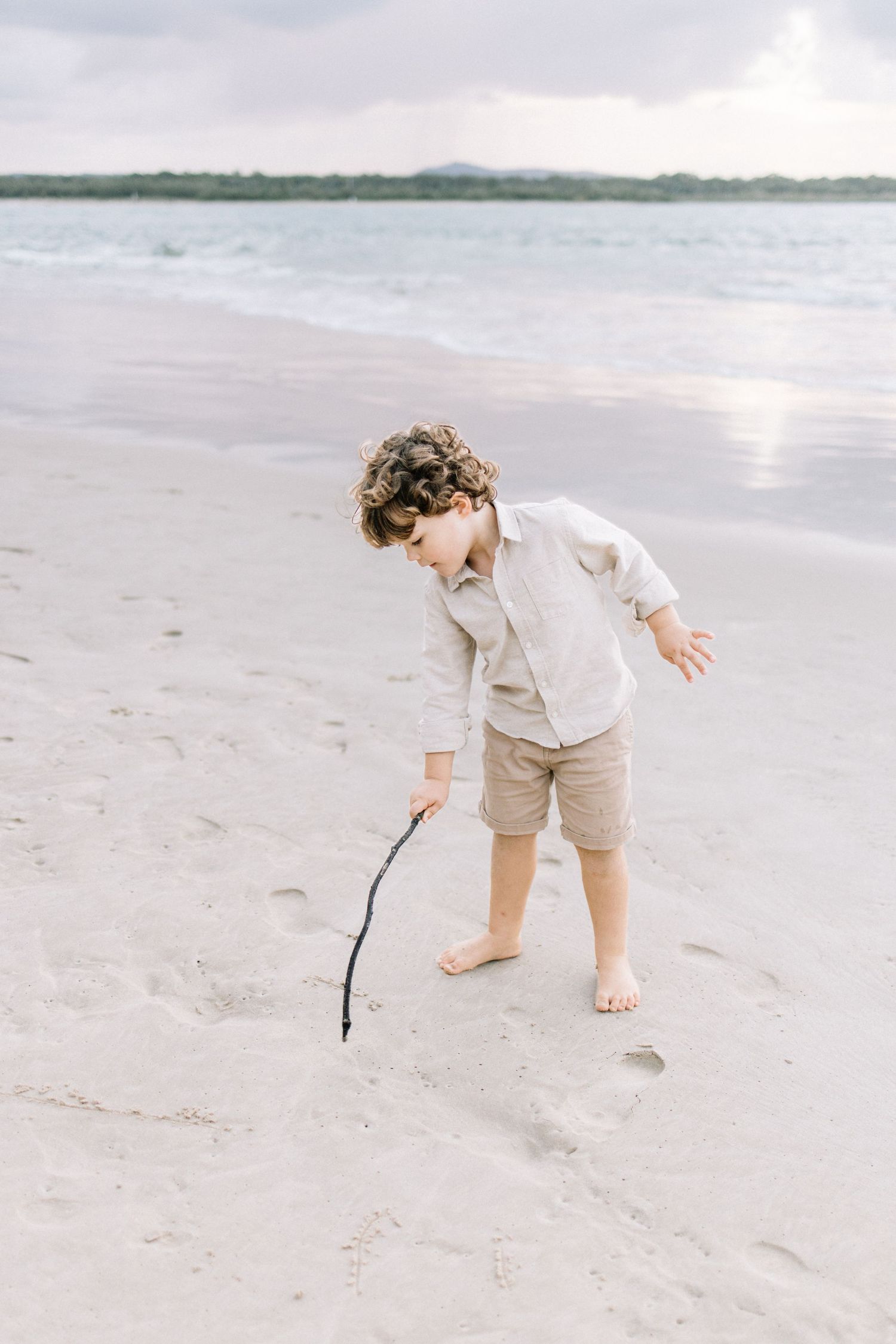 Curly-haired boy in linen shirt and shorts drawing in white sand with a stick on a serene beach.