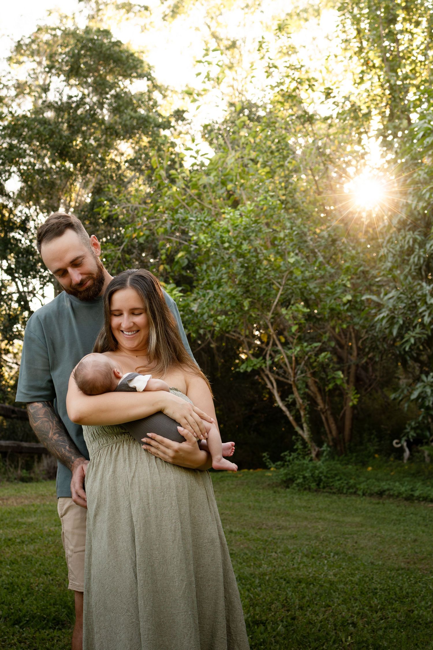 A couple stands in a sunlit garden, smiling at their newborn baby held tenderly in the mother's arms.