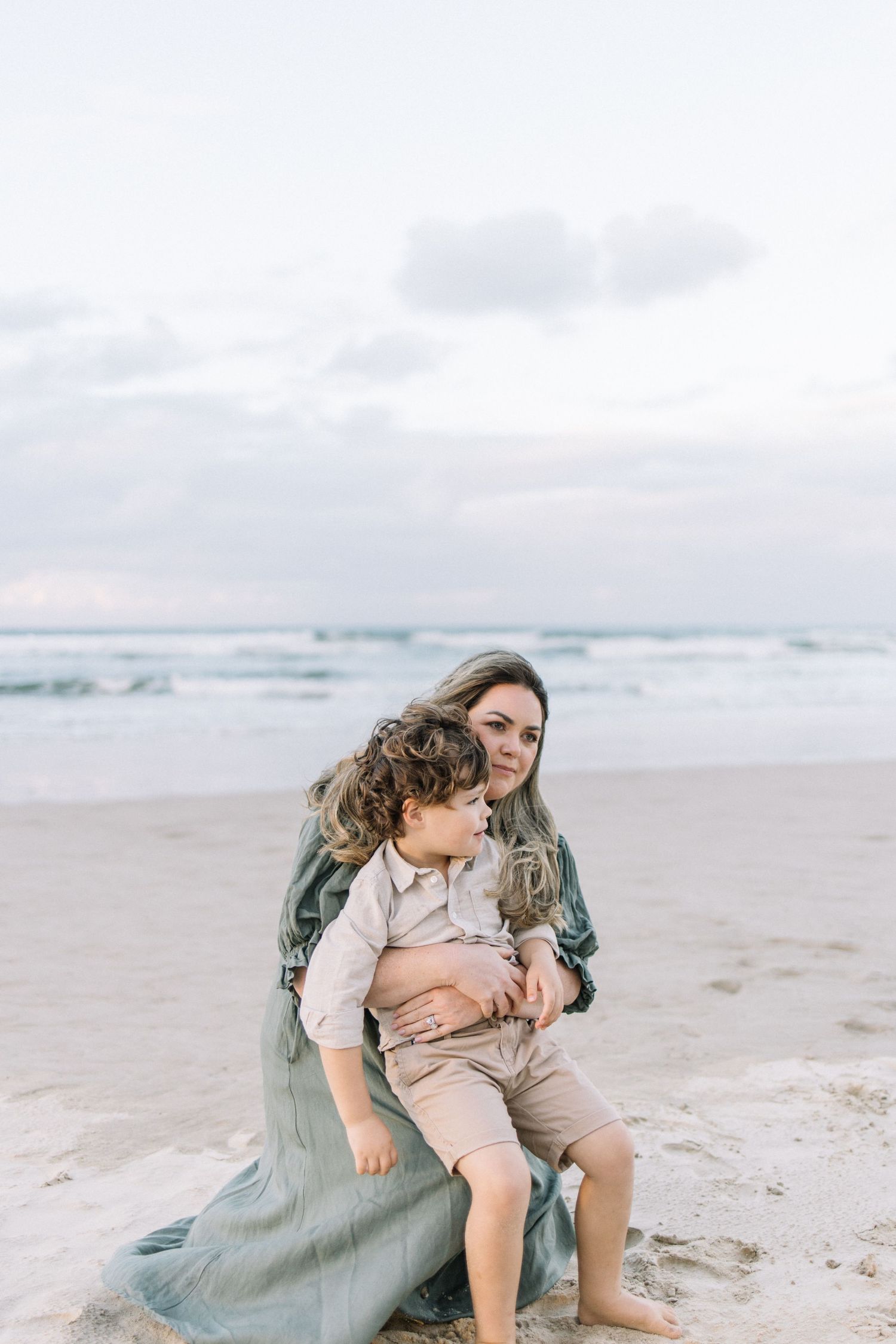 A mother in a flowing green dress sits on the beach cradling her curly-haired toddler son.