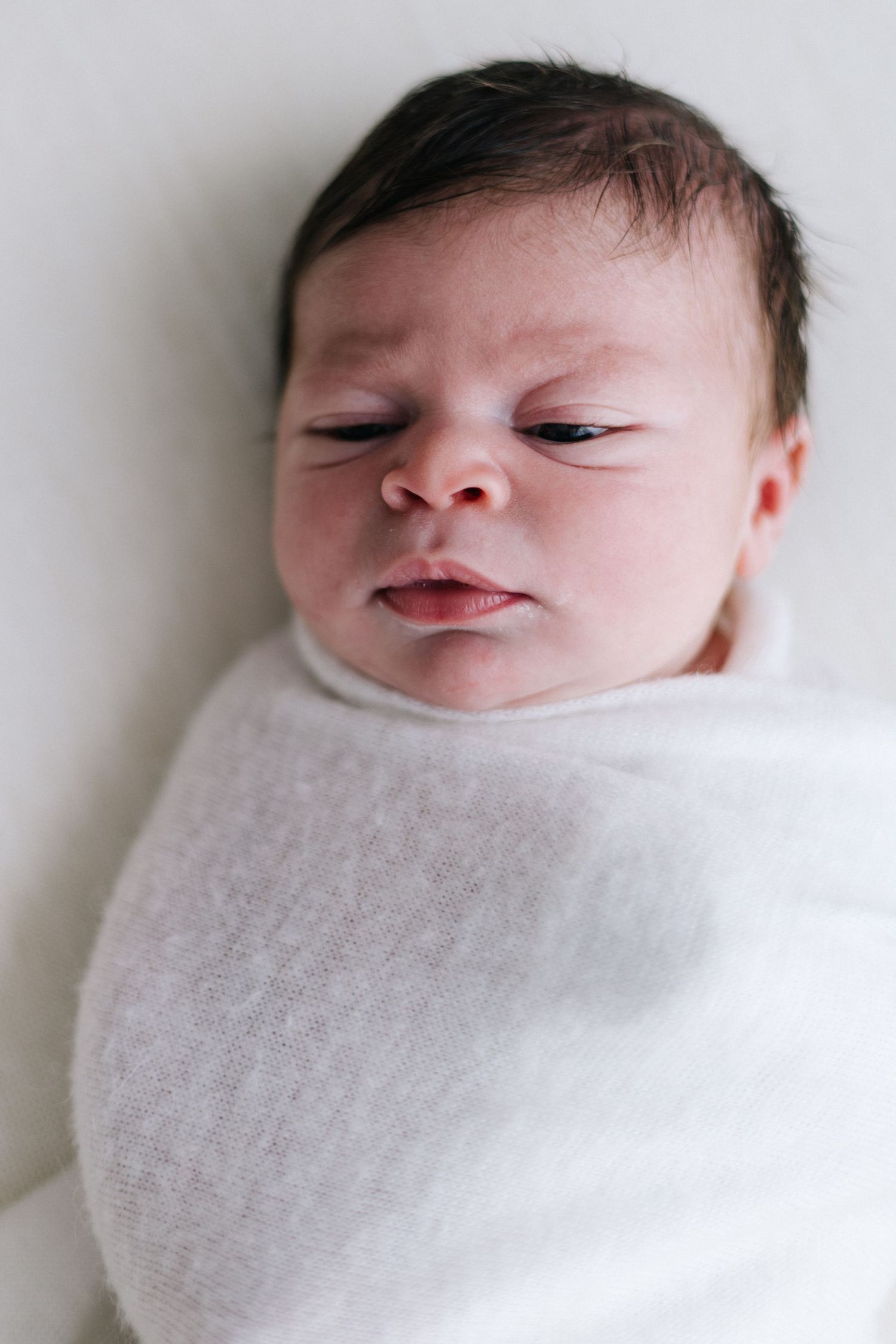 Newborn baby swaddled in white wrap with eyes closed, peaceful expression, dark hair visible.
