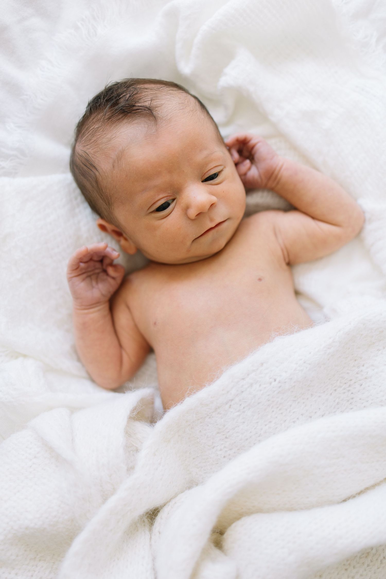 Newborn baby lying on a soft white blanket, with dark hair and tiny hands raised beside their head.
