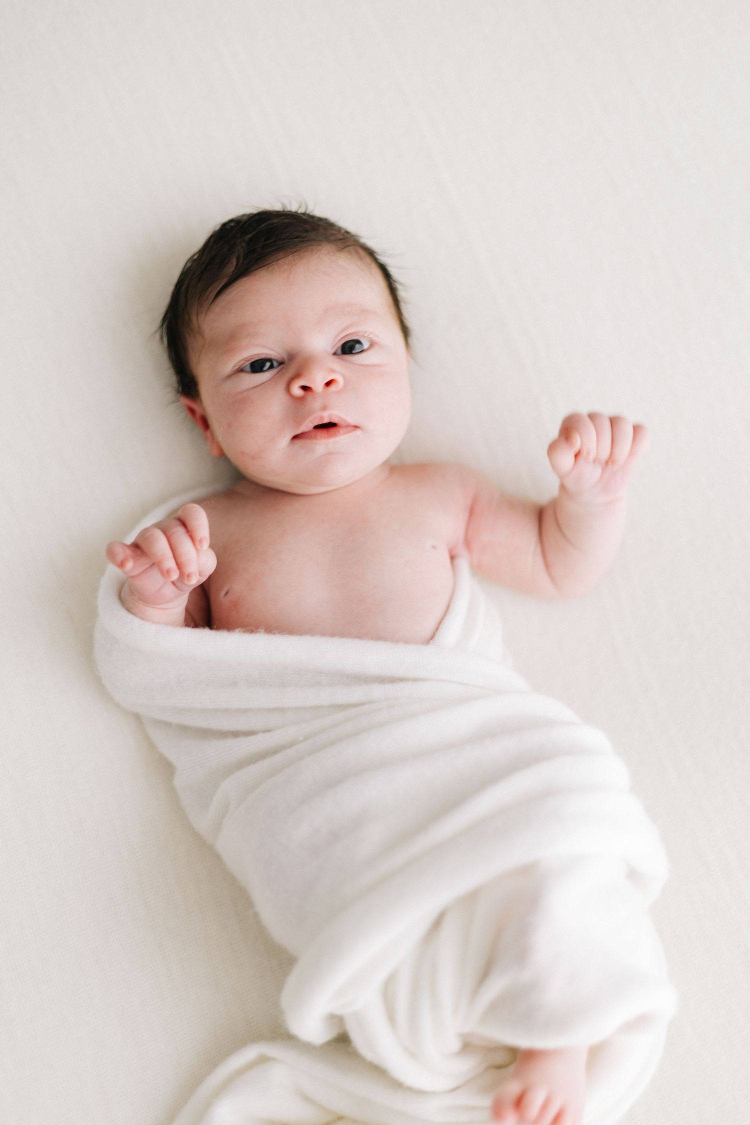 Newborn baby wrapped in a white swaddle blanket lying on a white surface with arms raised.