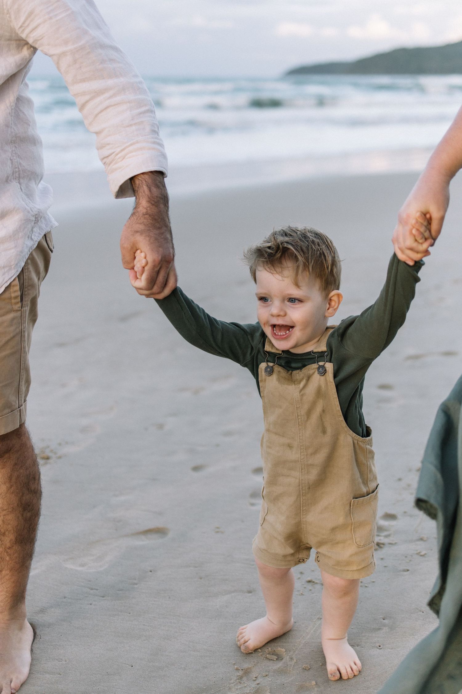 Joyful toddler boy holds hands with parents while standing barefoot on a sandy beach at dusk.