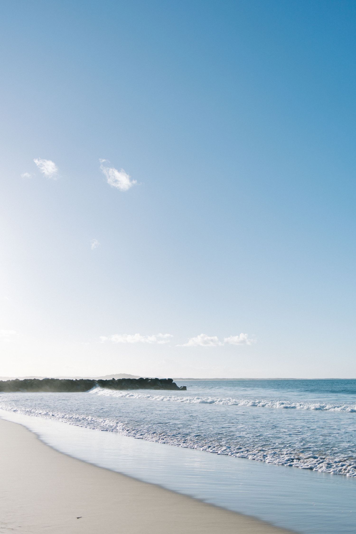 Serene tropical beach with gentle waves, white sand shoreline, rocky outcrop, and clear blue sky with wispy clouds.