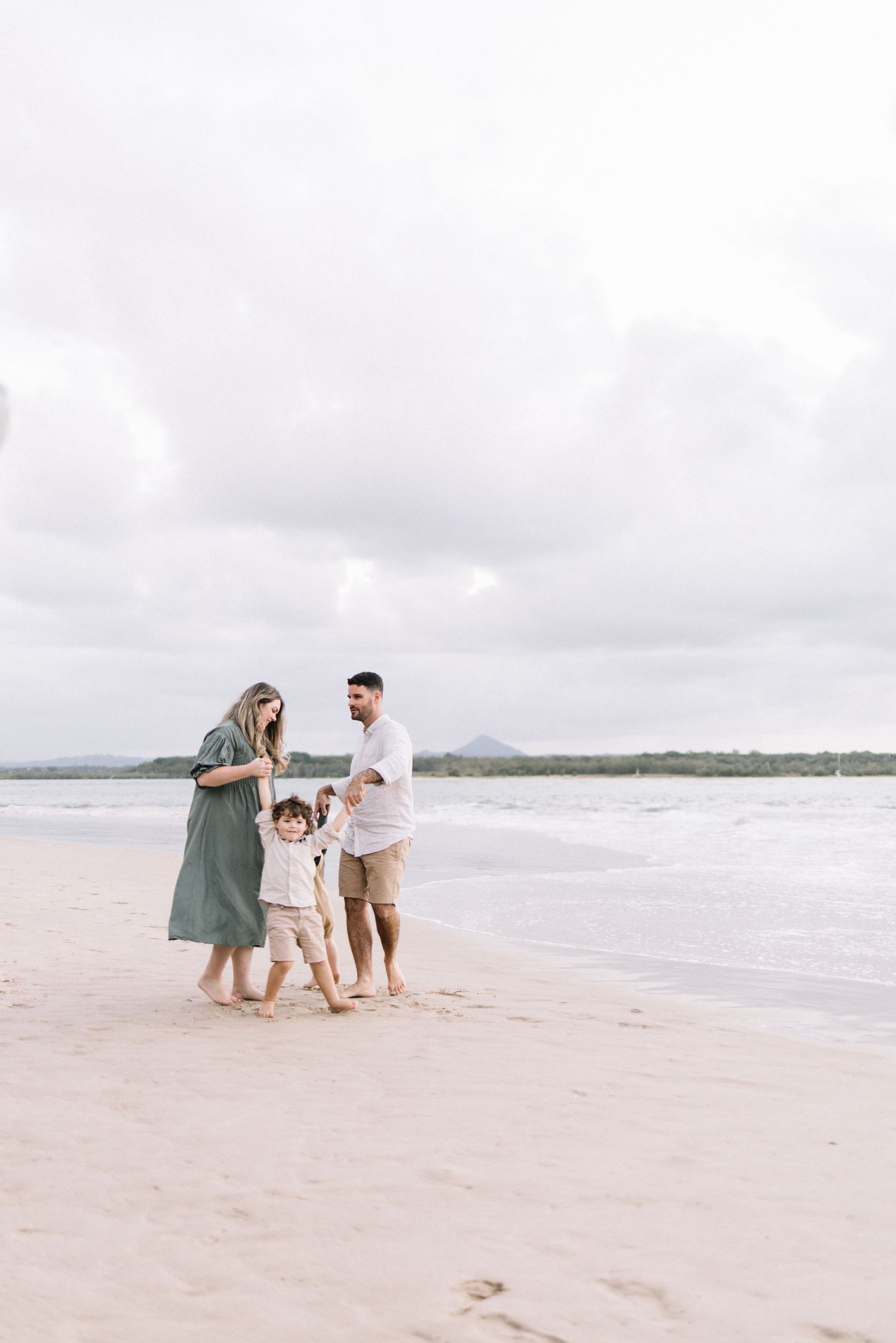 Young family of four walking hand-in-hand along a sandy beach with ocean waves and cloudy sky behind them.
