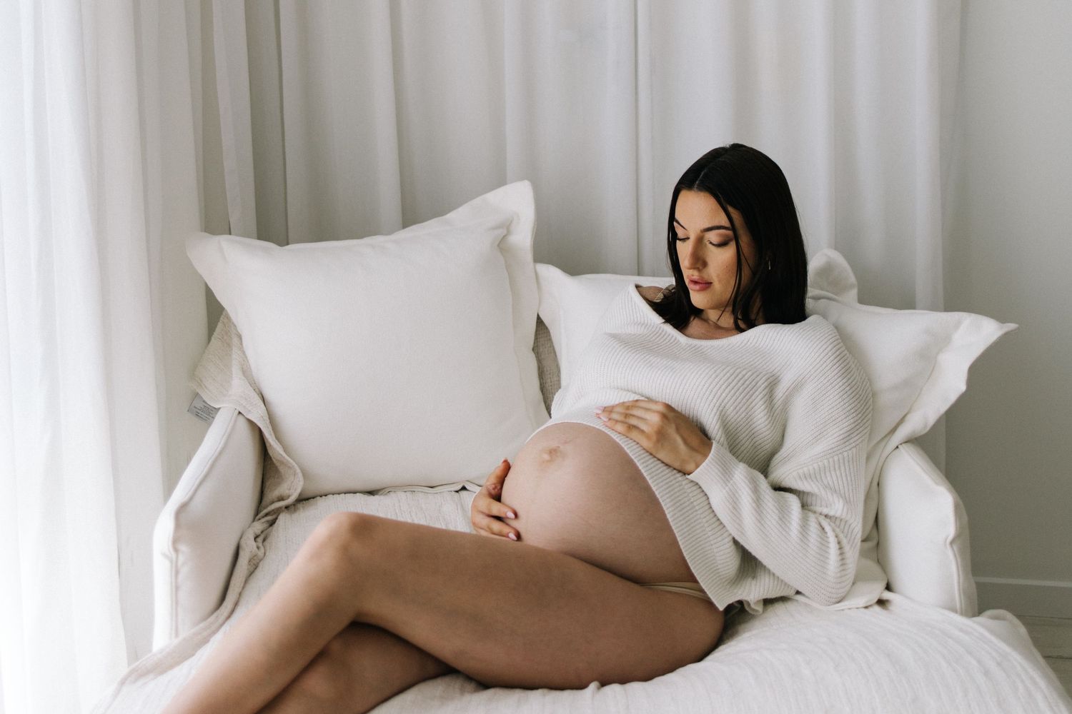 Pregnant woman in white sweater sitting on a chair, cradling her baby bump with both hands.