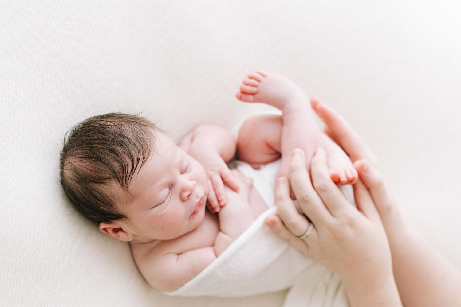 Peaceful newborn baby sleeping wrapped in white, cradled gently by mother's hands on soft white background.