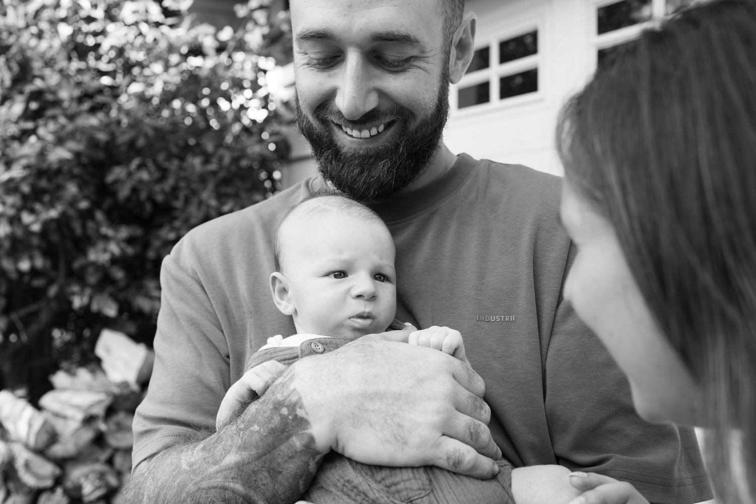 Bearded father smiling while holding newborn baby outdoors in black and white family portrait.