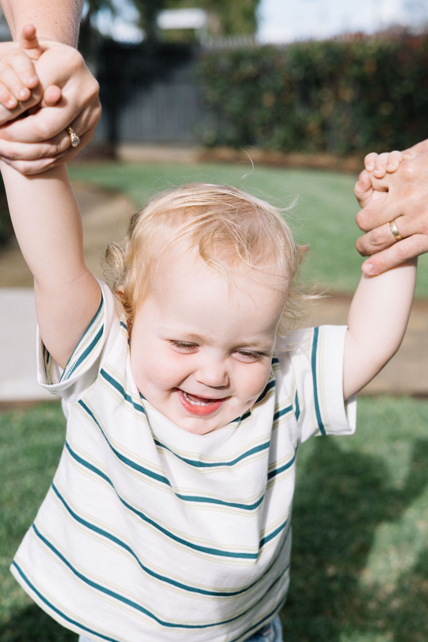 Happy toddler in striped shirt laughing while being lifted by hands outdoors on sunny day.