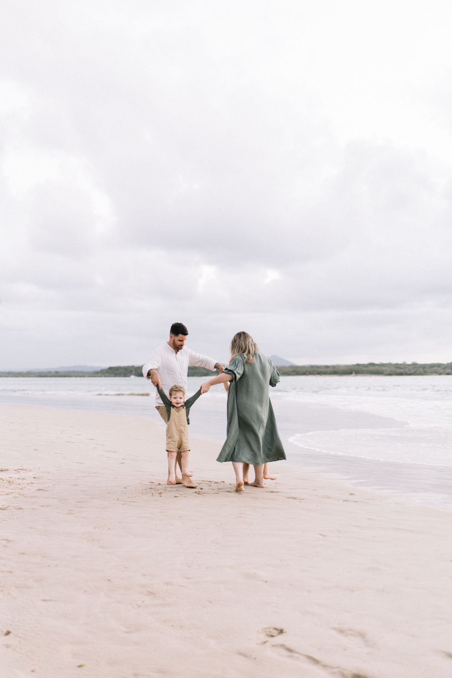 Parents in neutral tones swing their young child by the hands on a cloudy beach.