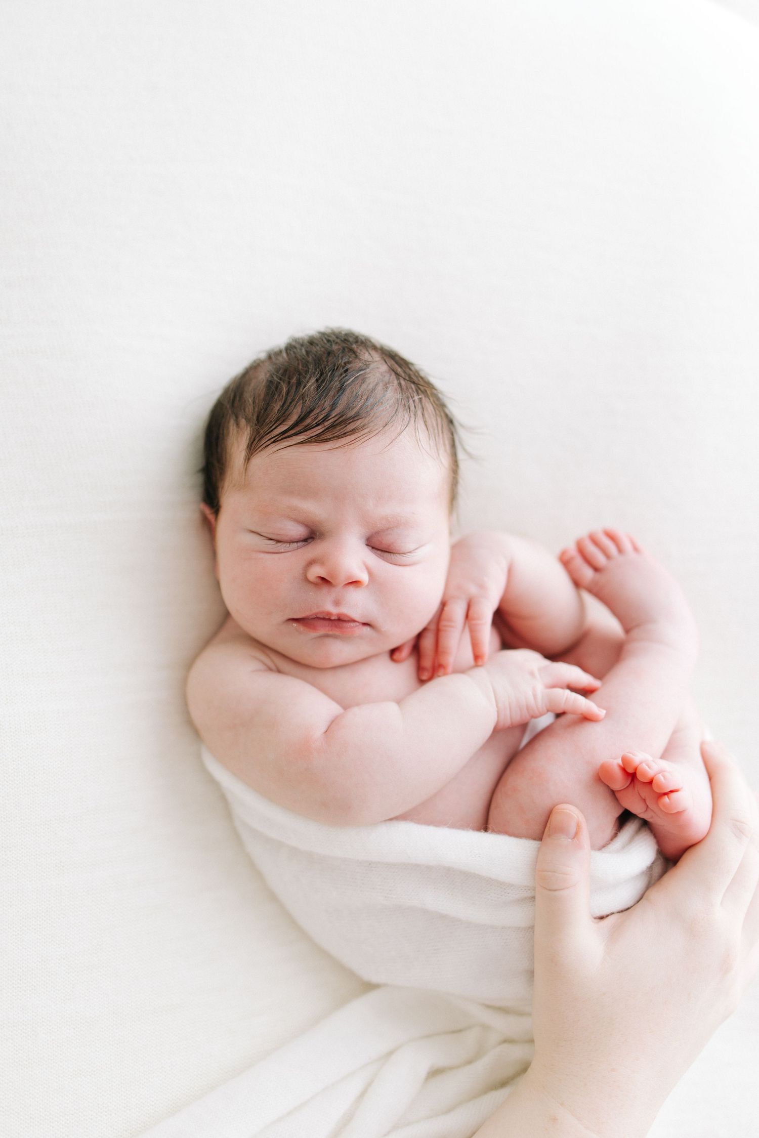 Peaceful sleeping newborn loosely swaddled in white with adult hand nearby.